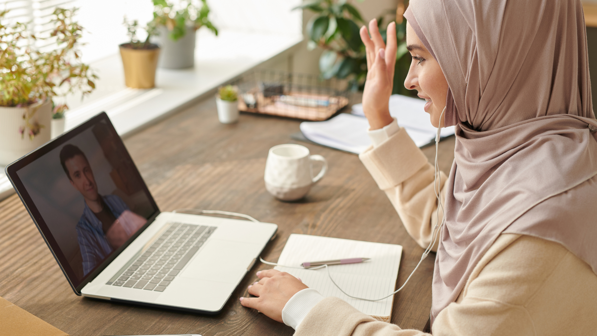 A woman wearing a hijab participates in a video call on her laptop, smiling and waving with her right hand, seated at a wooden table with notebooks, a pen, and a mug, with plants on the windowsill in the background.