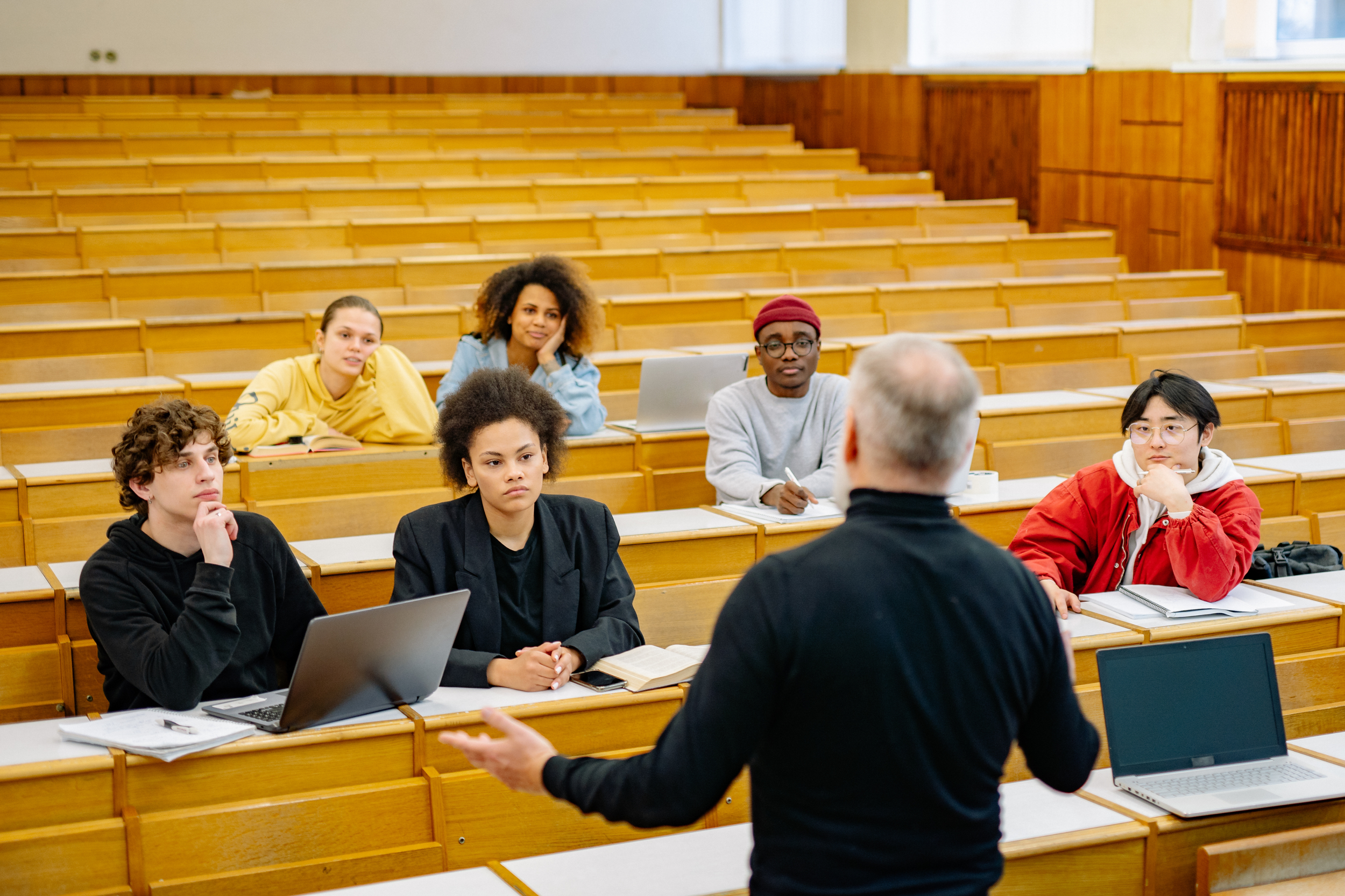 Professor speaking to students in an empty classroom with wooden desks and laptops.