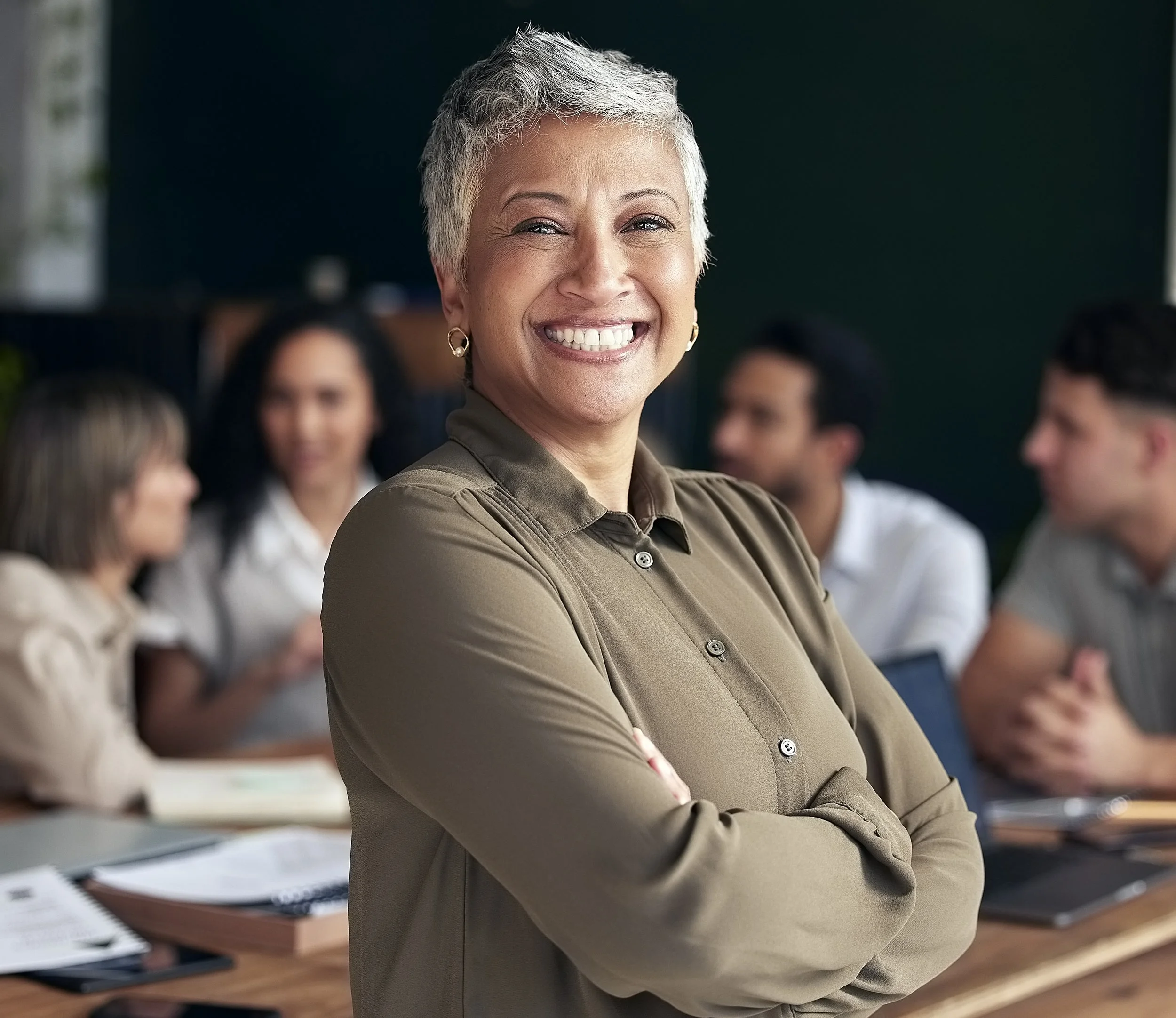 Smiling woman with short gray hair and earrings standing with arms crossed in front of a group of people in a meeting room.