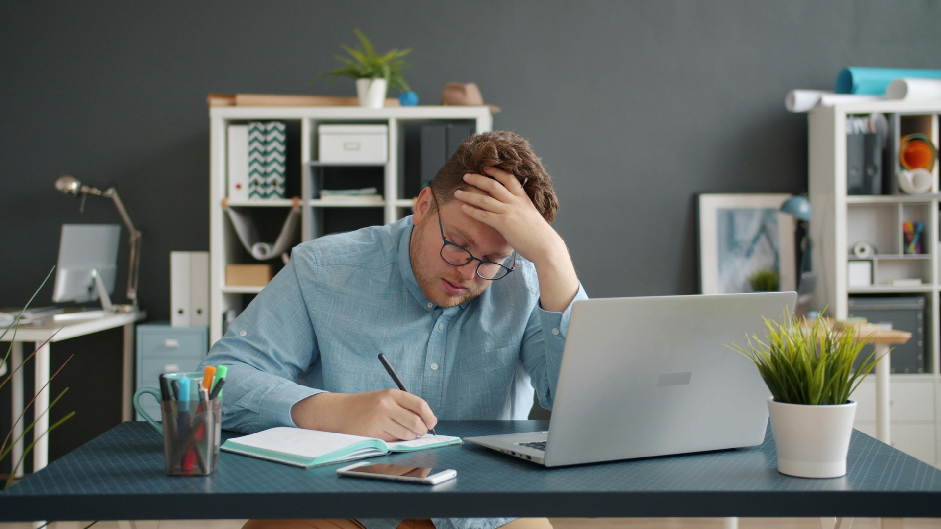 A man sitting at a desk with a laptop, holding his forehead in frustration, taking notes in a notebook, with a smartphone and a cup of pens on the desk, in a modern office with shelves and plants.