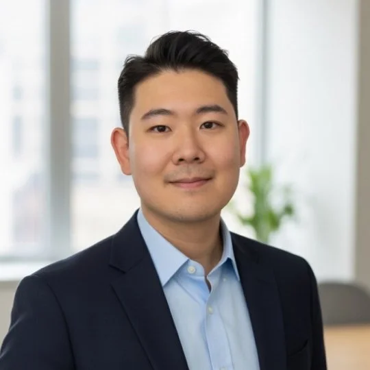 Professional man in a suit smiling in an office setting with large windows and a plant in the background.