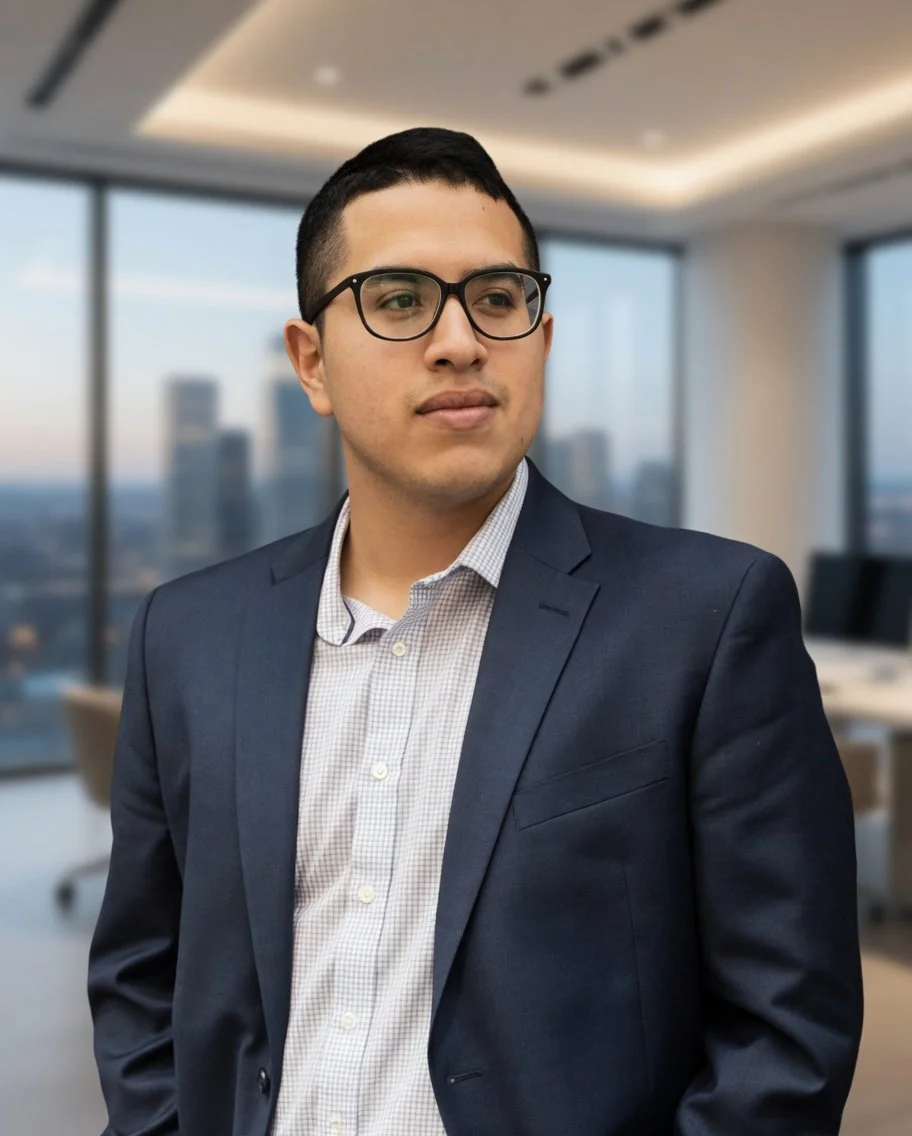 A young man with dark hair, glasses, and light skin wearing a navy blazer and a white checkered shirt, standing in a modern office with large windows showing a city skyline in the background.
