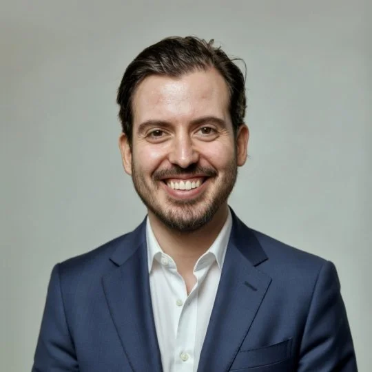 A man with dark hair and a beard wearing a navy blue suit and white shirt, smiling against a plain light gray background.