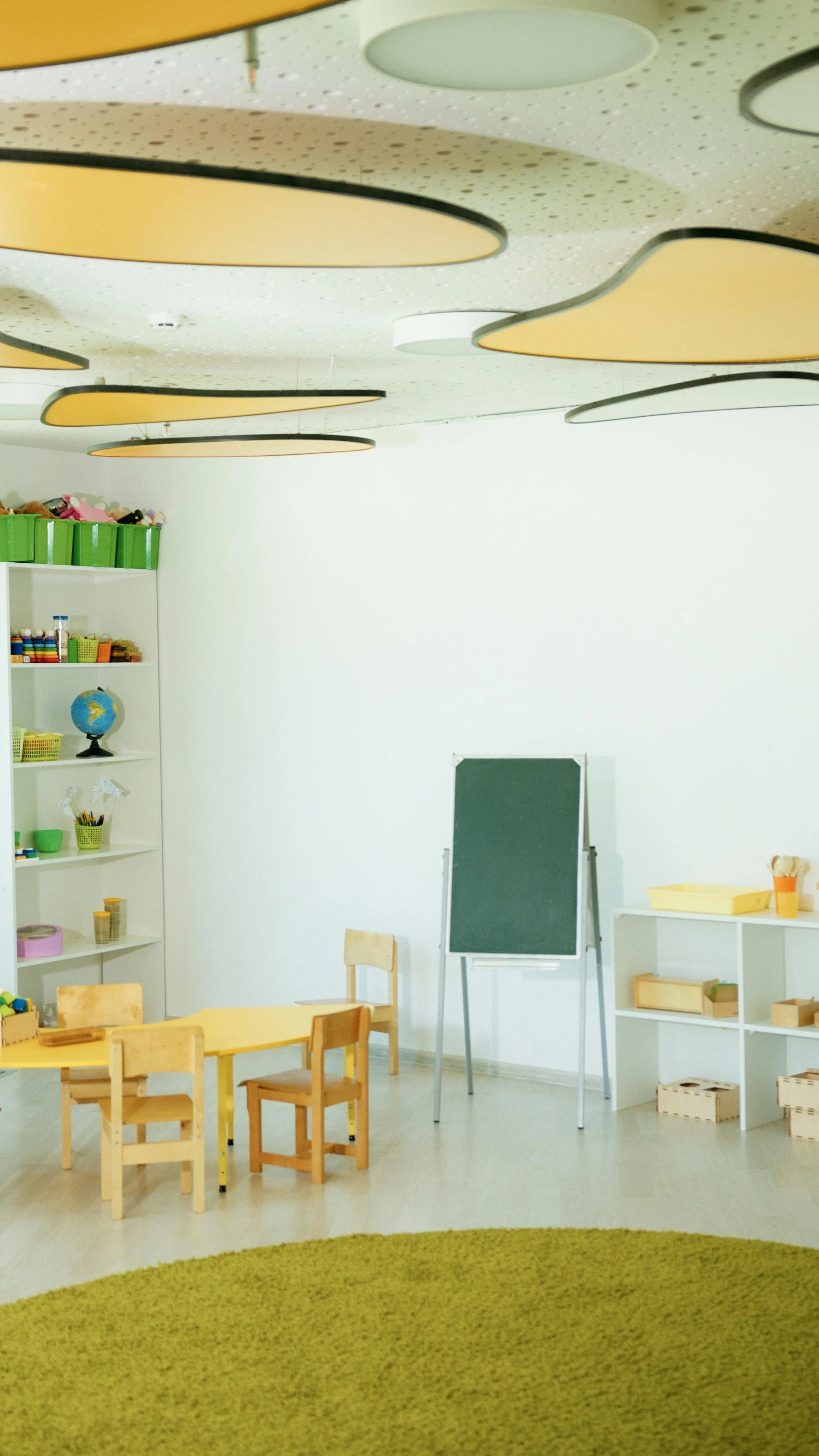 Empty classroom with small wooden tables and chairs, a chalkboard on a stand, white shelves with educational supplies, a green rug, and ceiling lights.
