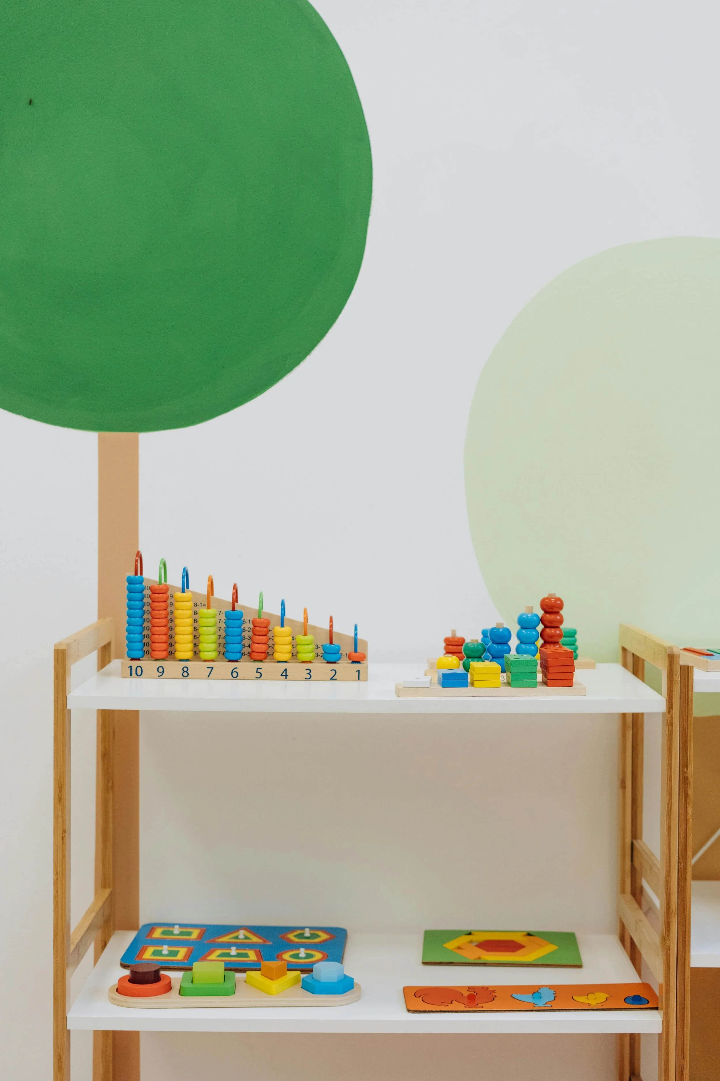 Children's educational toys on a white and wooden shelf, including stacking rings, geometric blocks, and shape sorters in a classroom or playroom setting.