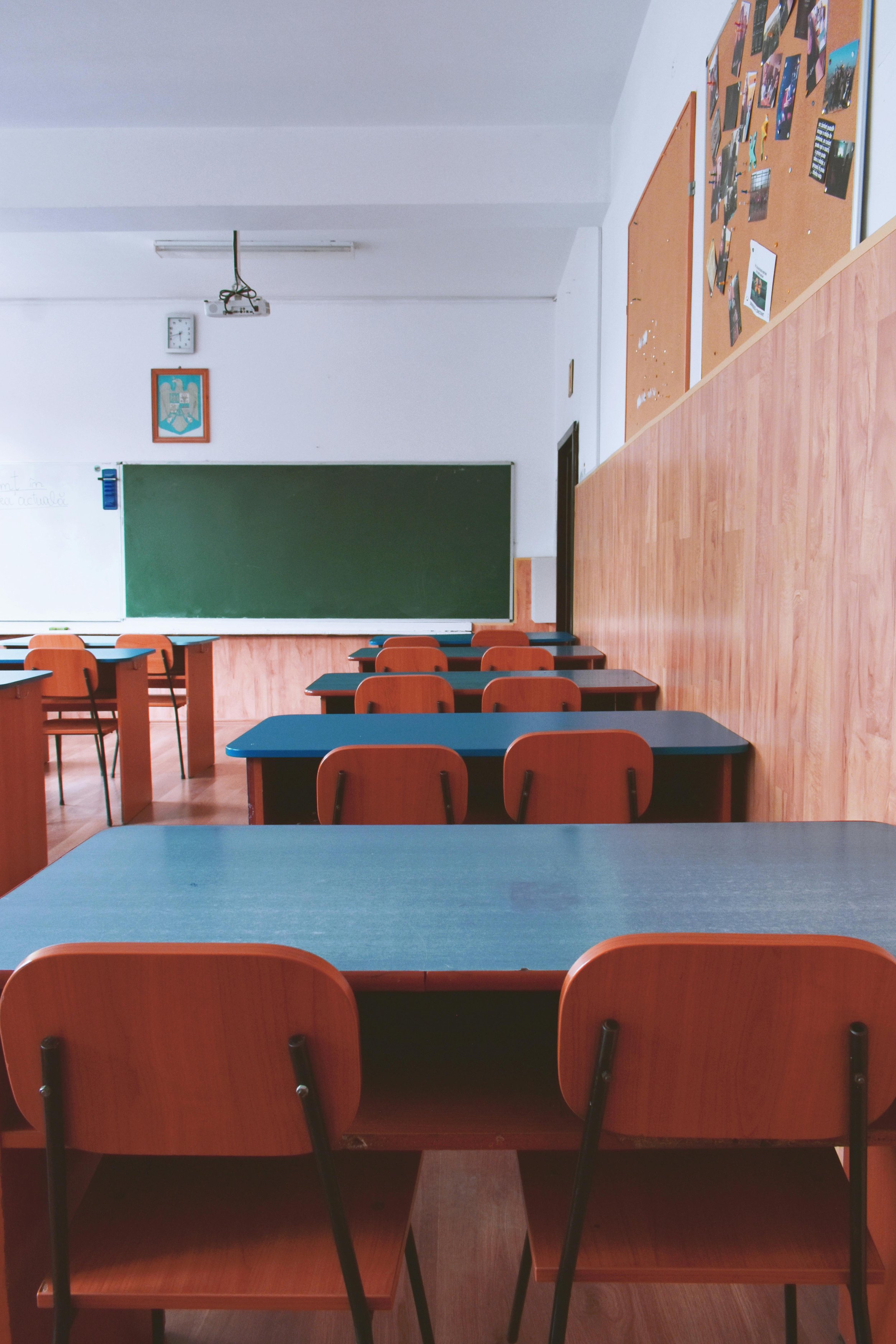 An empty classroom with desks and chairs facing a green chalkboard on the wall. There is a bulletin board with photos and papers on the right side of the room, and a picture of a coat of arms hung above the chalkboard.