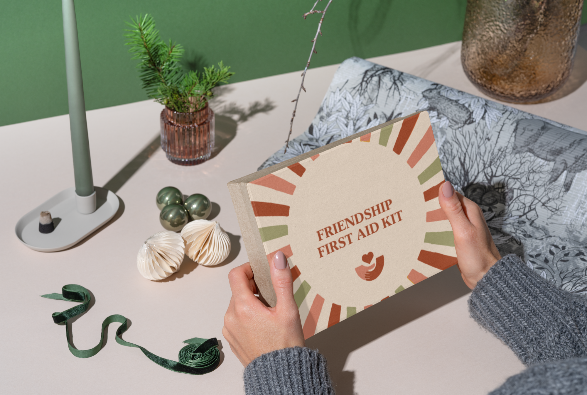 Person holding a box labeled 'Friendship First Aid Kit' at a table decorated with a small potted plant, metallic ornaments, foldable paper decorations, and a green ribbon.