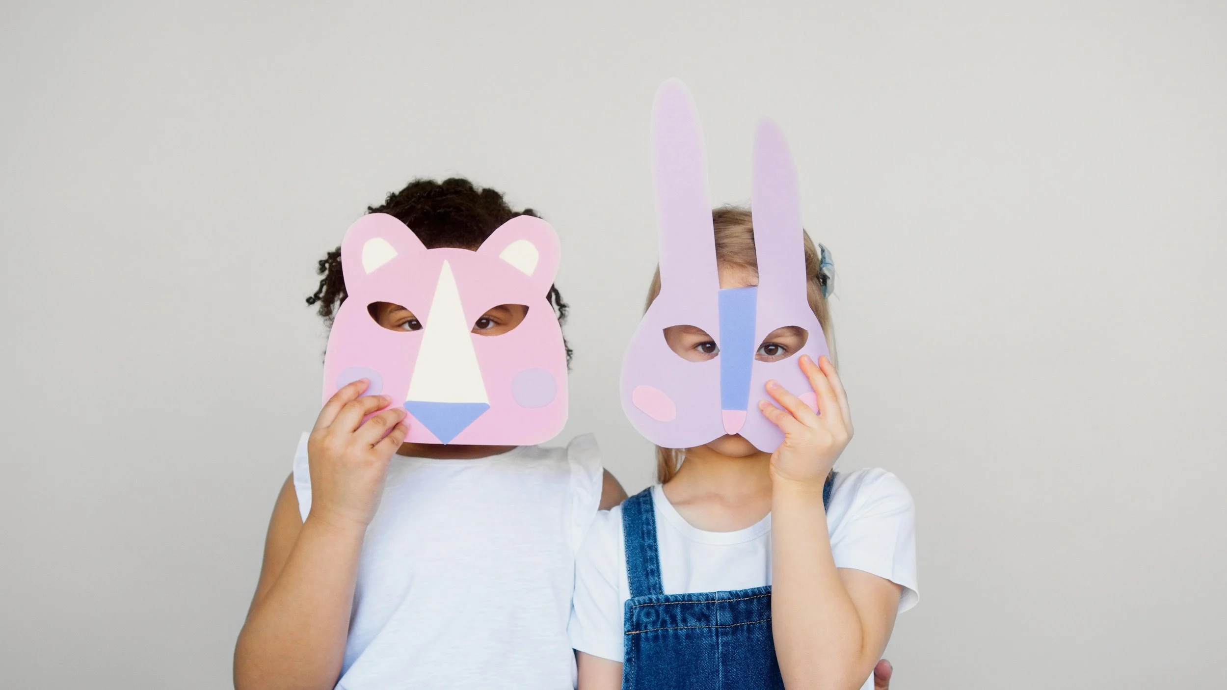 Two children wearing animal masks, one with a bear mask and the other with a rabbit mask, standing against a plain background.