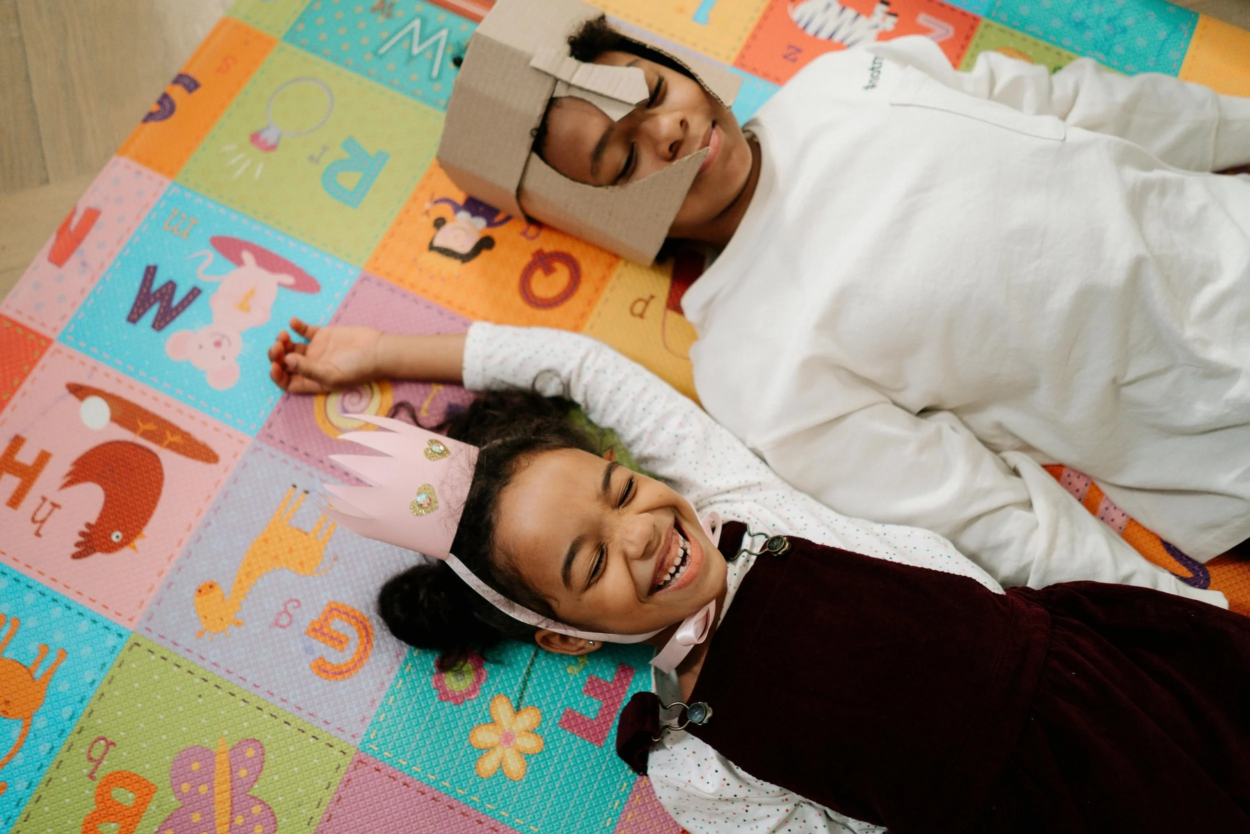 A young girl wearing a pink birthday crown, smiling happily, lying on a colorful alphabet quilt with a big smile. Beside her, a person with short hair, closed eyes, and a cardboard crown on their head, also lying down. They are holding hands.