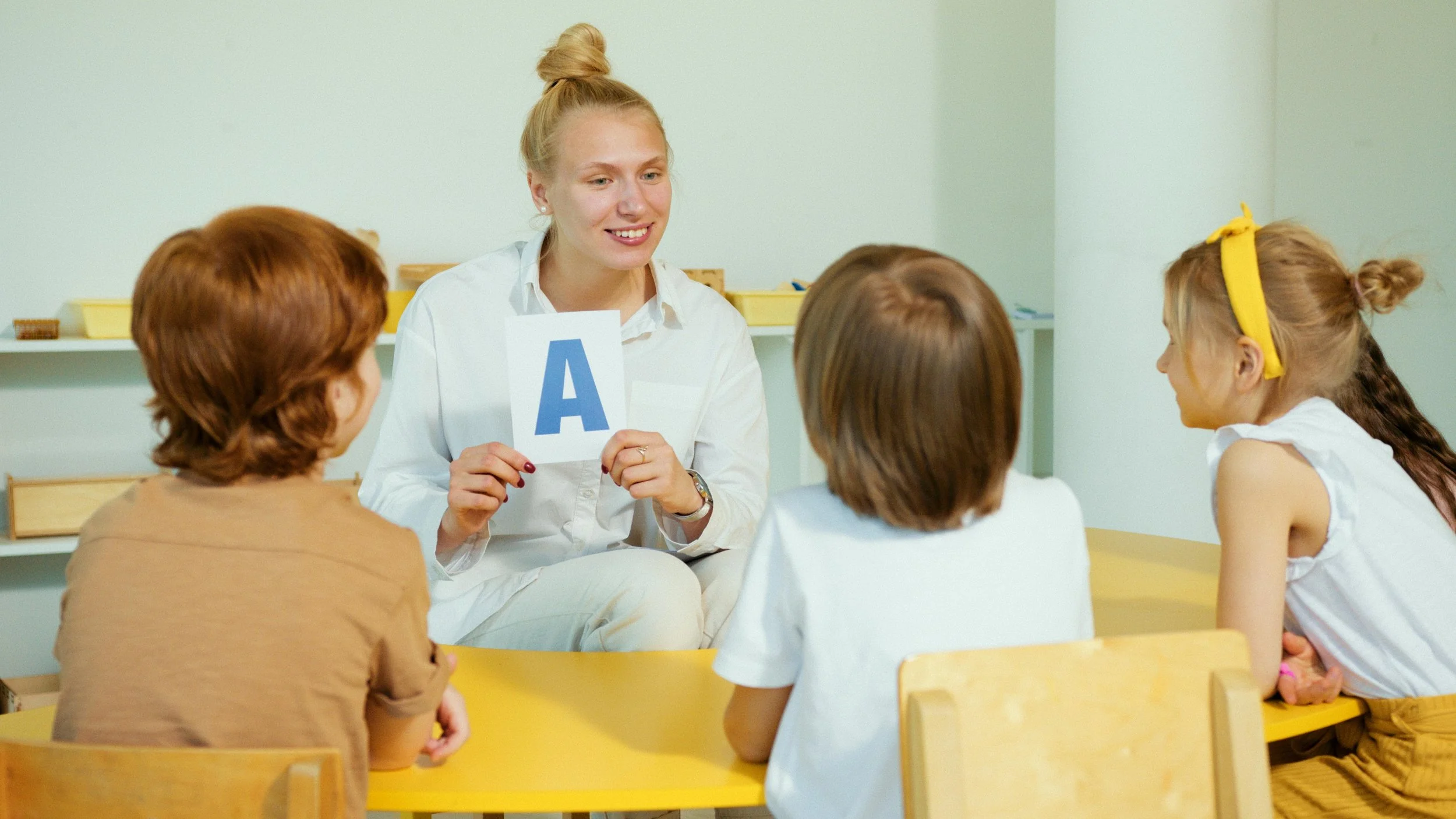 A teacher holding a flashcard with the letter A on it, engaging with a group of four children at a yellow table in a classroom.