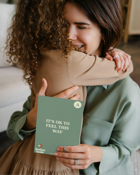 Two women hugging indoors, one holding a green book that reads 'It's OK to Feel This Way' and 'Fearless Inside'.