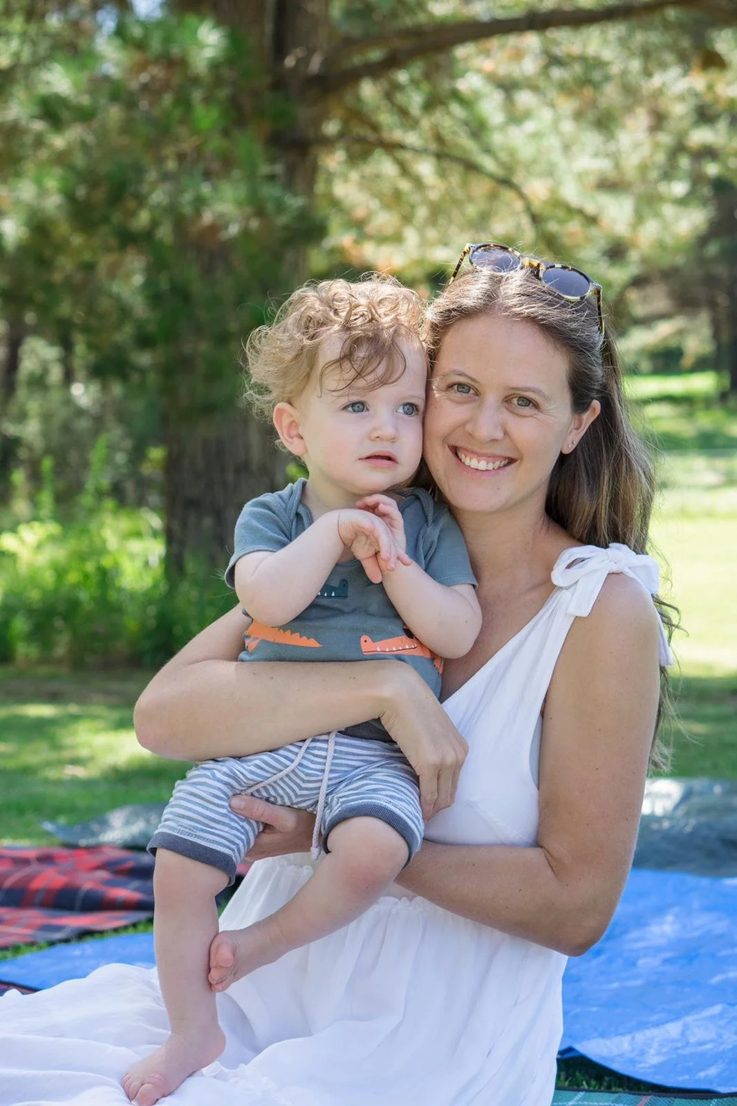 A woman holding a young boy outdoors on a sunny day. They are in a green park with trees in the background. The woman is smiling, wearing a white dress and sunglasses on her head. The boy has curly blonde hair, blue eyes, and is wearing a gray shirt with dinosaur prints and striped shorts.