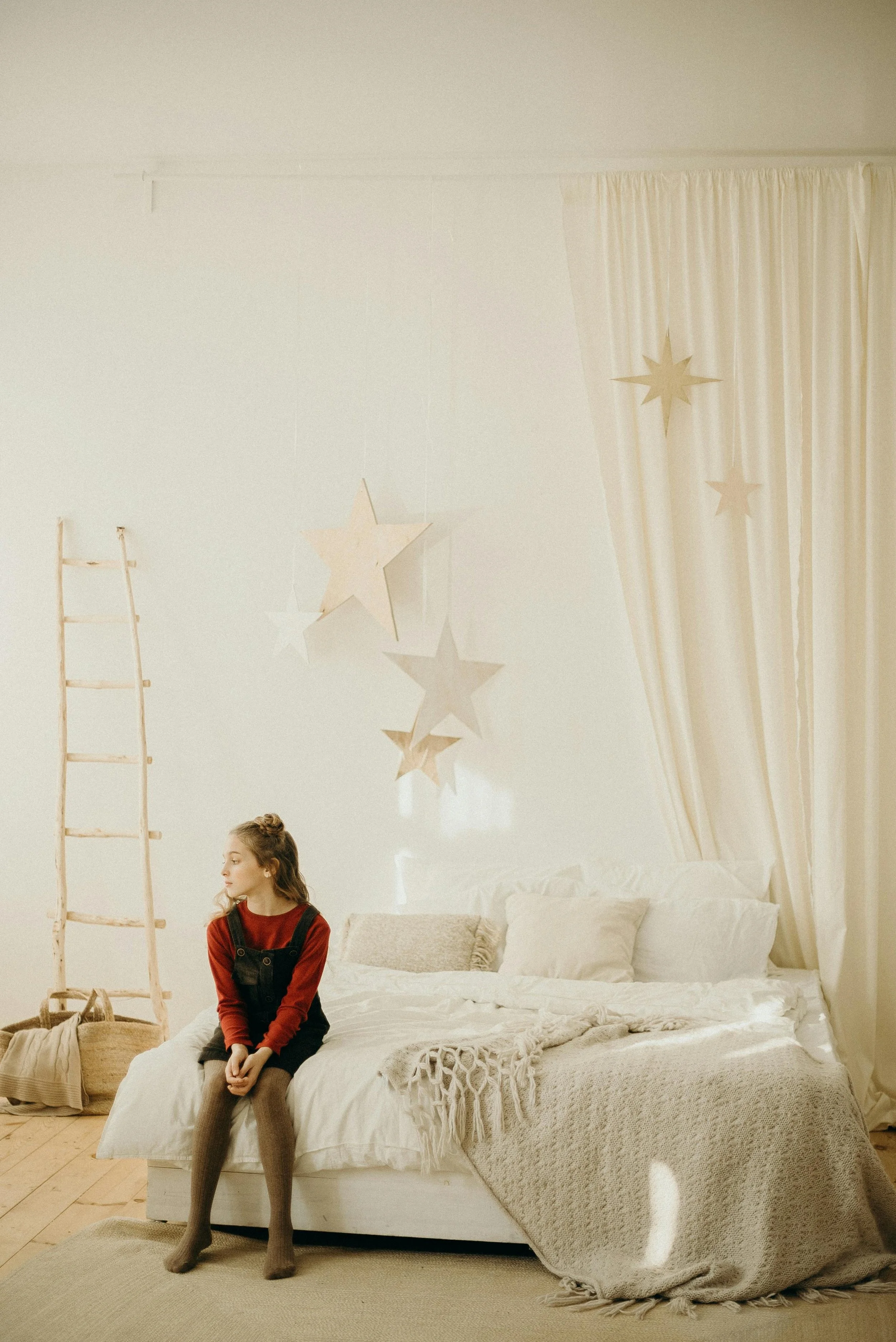 A girl sitting on a bed in a cozy, neutral-toned bedroom with star-shaped hanging decorations, a ladder-style decor piece, and soft blankets and pillows.
