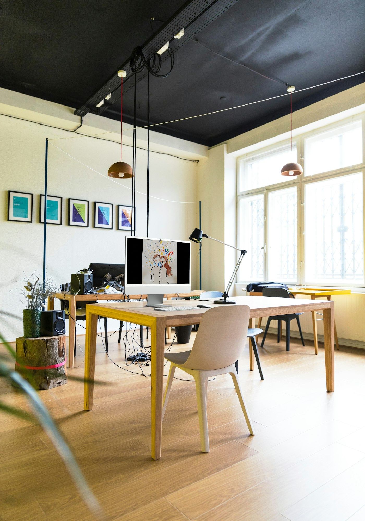 A bright, modern office with wooden floors and large windows. Two desks with computers, chairs, and various office supplies. Decorative framed artworks on the wall, a plant, and hanging pendant lights.