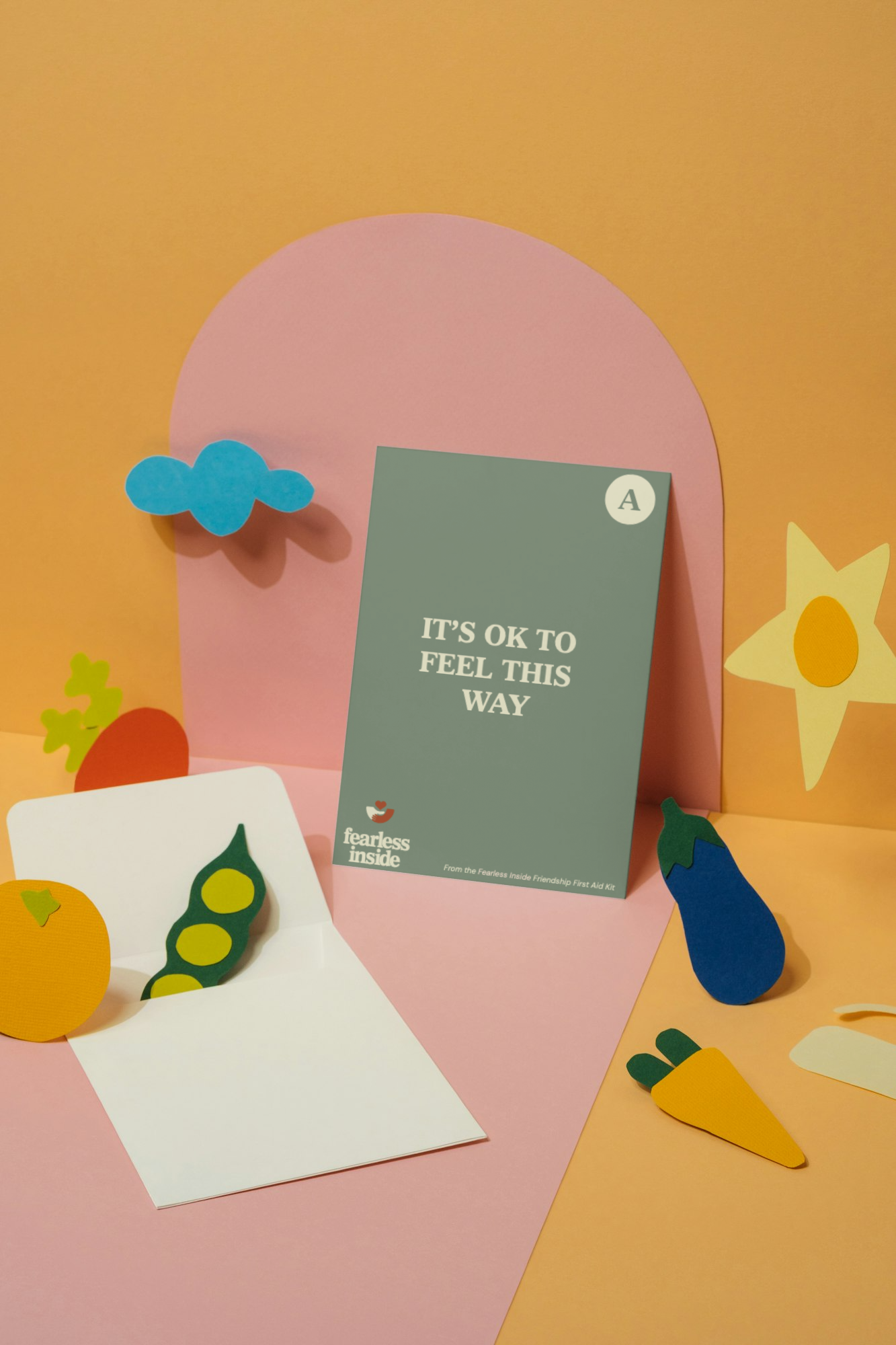 A pink and orange background with paper cutouts of a blue cloud, a yellow star, a carrot, an eggplant, a yellow tomato, and a white notecard with a green pea pod. A gray card with white text that reads 'IT'S OK TO FEEL THIS WAY' is placed against a p