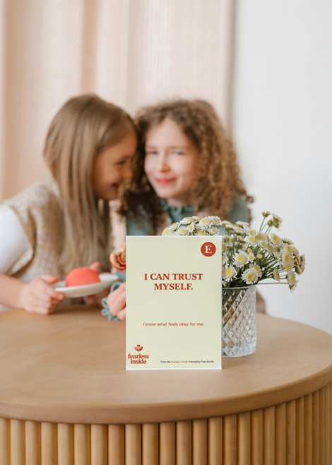 Two young girls with curly hair smiling near a table with a vase of white and yellow flowers and a card that says, 'I CAN TRUST MYSELF.'