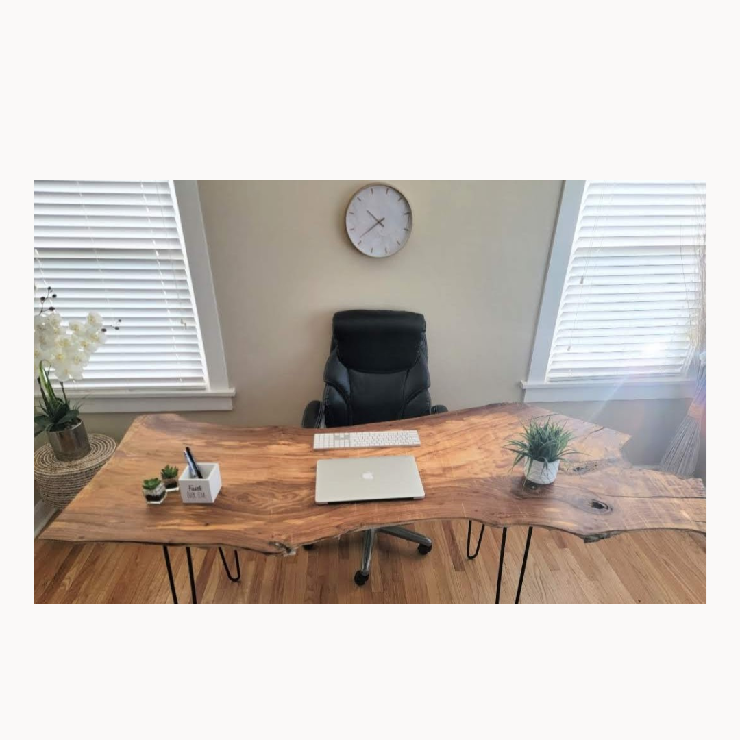 A contemporary office desk with a live edge wood top, black office chair, and decorative plants, with a wall clock and windows with blinds in the background.