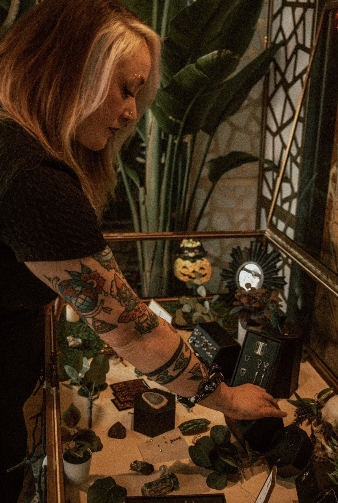 A woman with blonde hair and colorful tattoos on her arm is looking at jewelry and gemstone items on display inside a decorative glass case at an indoor event or shop, with plants and decorative items in the background.