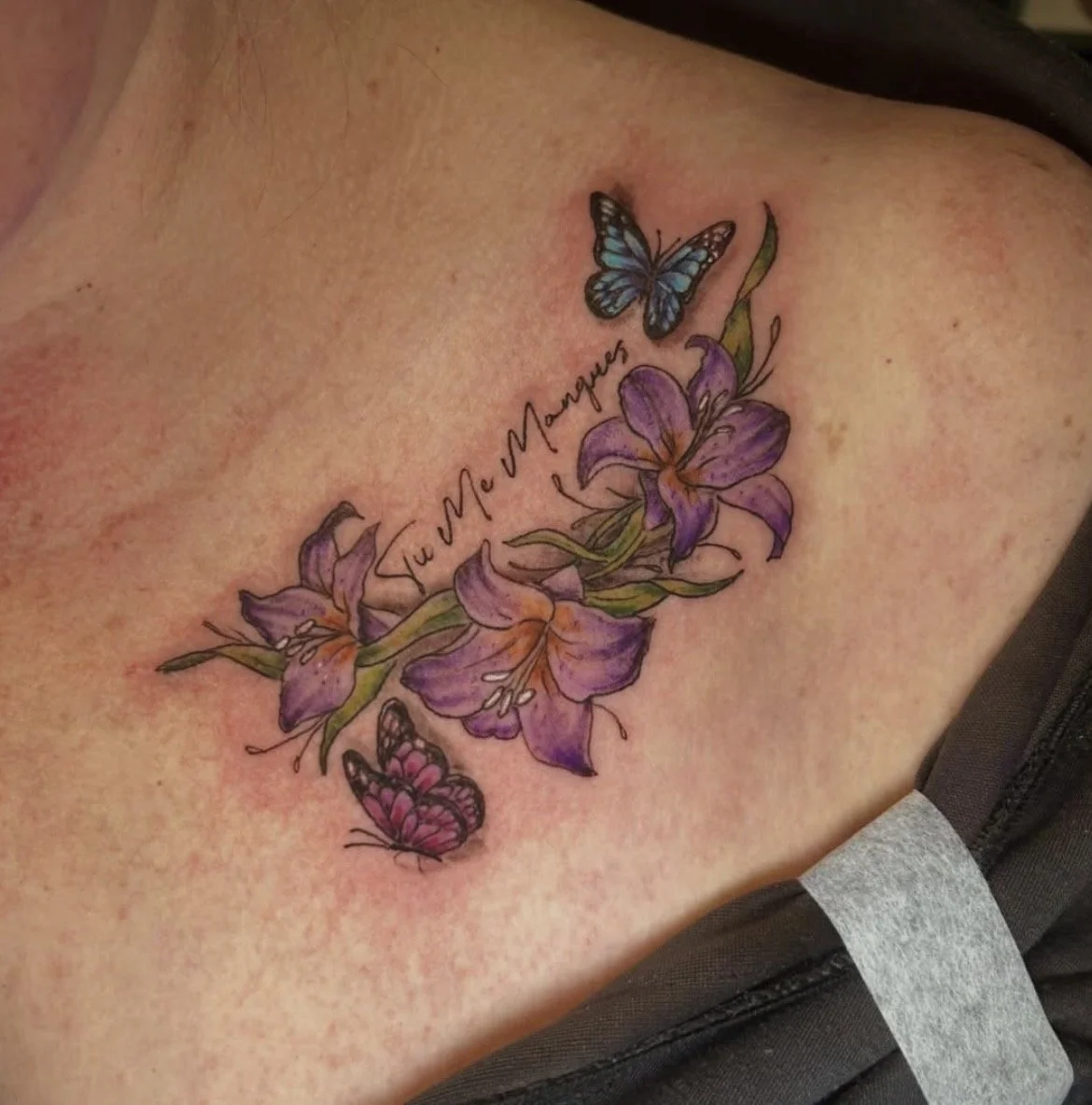 A colorful tattoo of purple lilies with green leaves and two butterflies, one blue and the other pink, along with the phrase 'Tu y Me Manques' written in cursive.