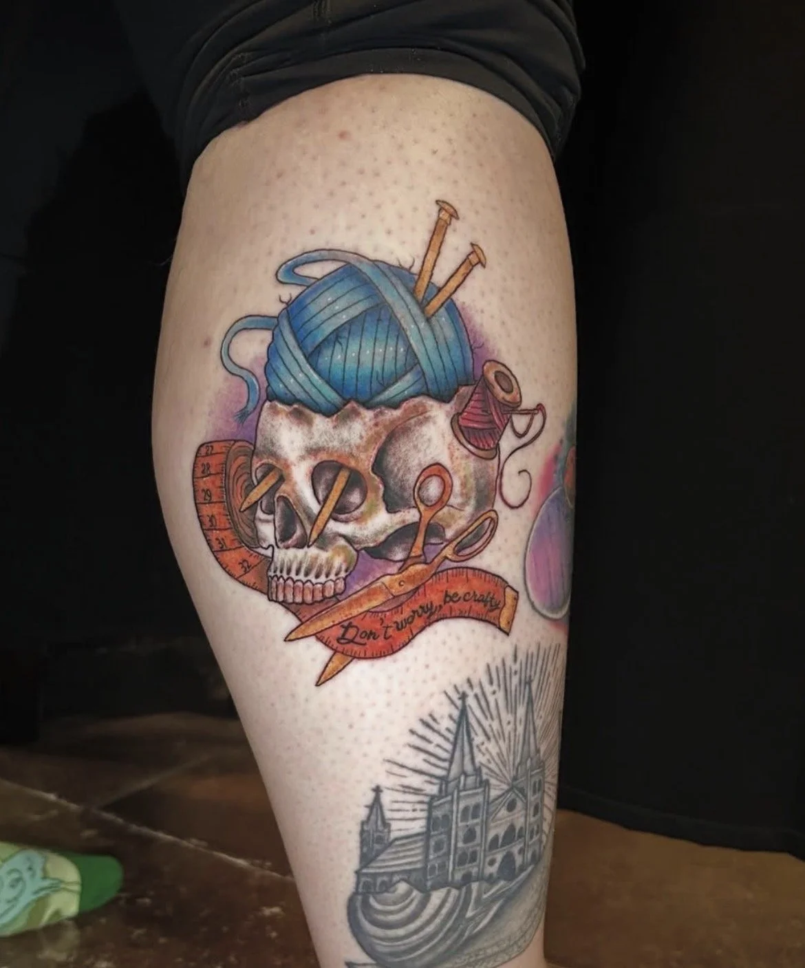 Tattoo of a skull with knitting yarn, scissors, and sewing accessories, with a quote ribbon and a church in the background.