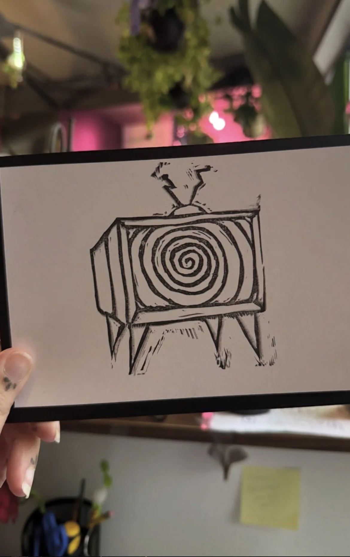 A hand holding a black and white drawing of a cassette tape with a spiral pattern on the label, standing on a small stand.