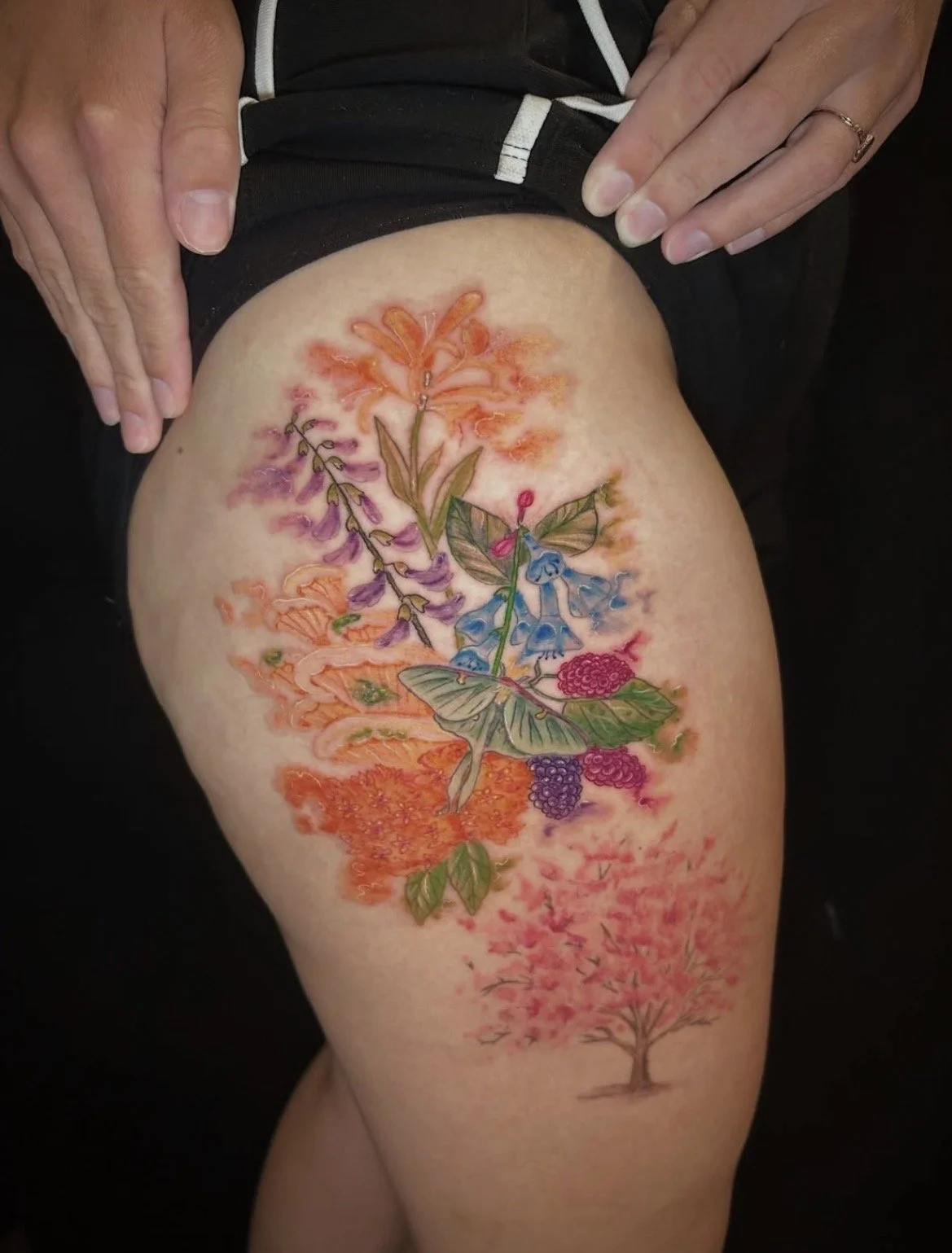 Colorful floral tattoo on person's thigh featuring various types of flowers, leaves, and a small tree.