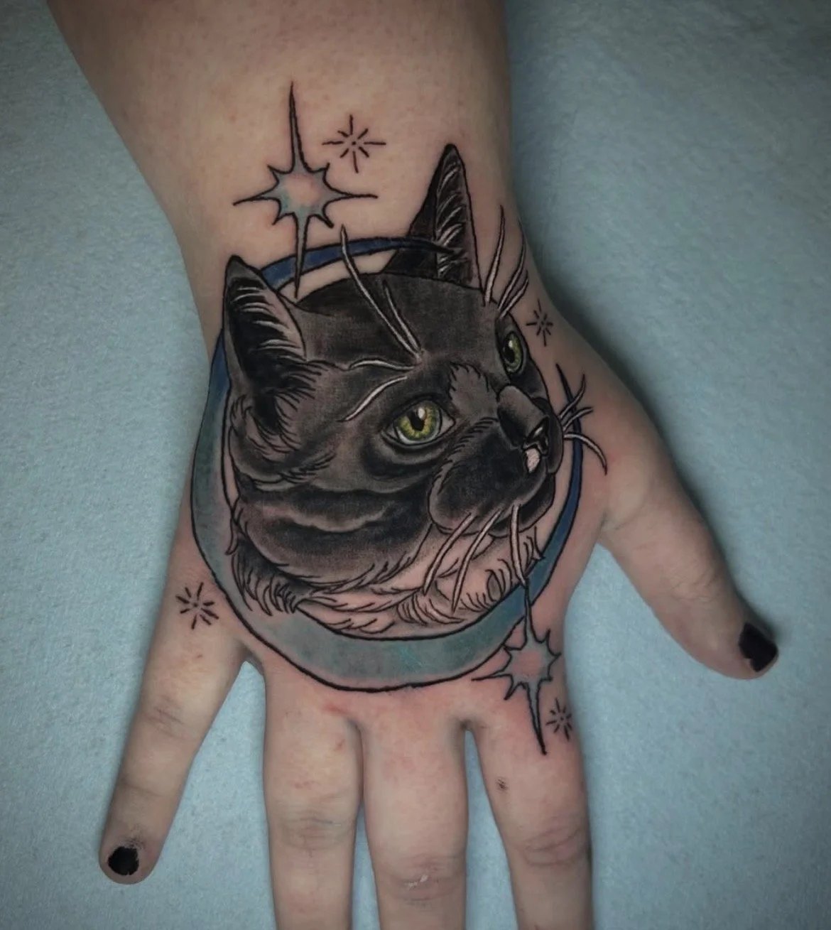 Tattoo of a cat's head inside a space helmet with stars and circles around it on a person's hand.