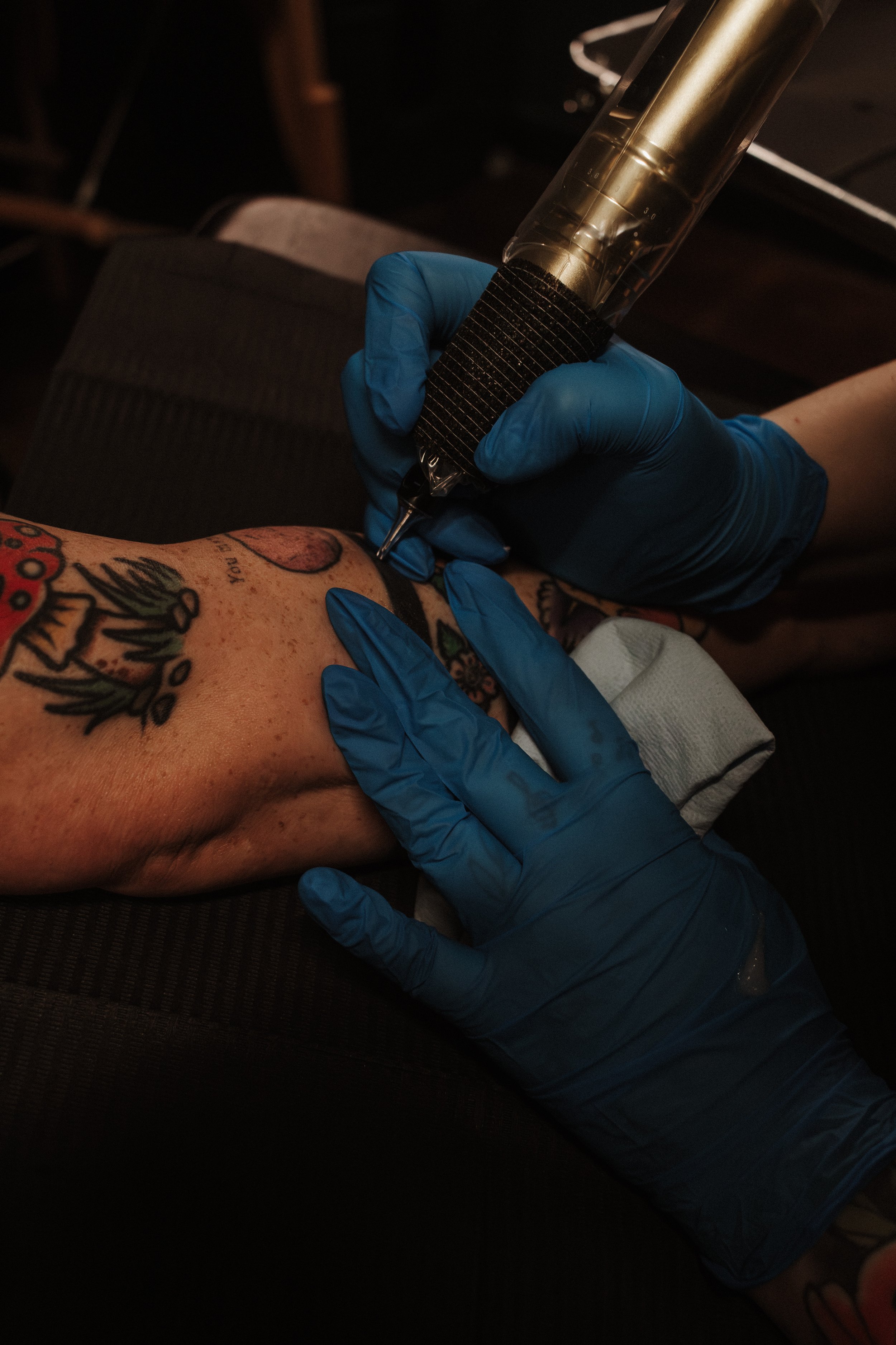A tattooed person's arm resting on a surface while a person wearing blue gloves uses a tattoo machine to create a tattoo on their forearm outdoors in a dimly lit setting.