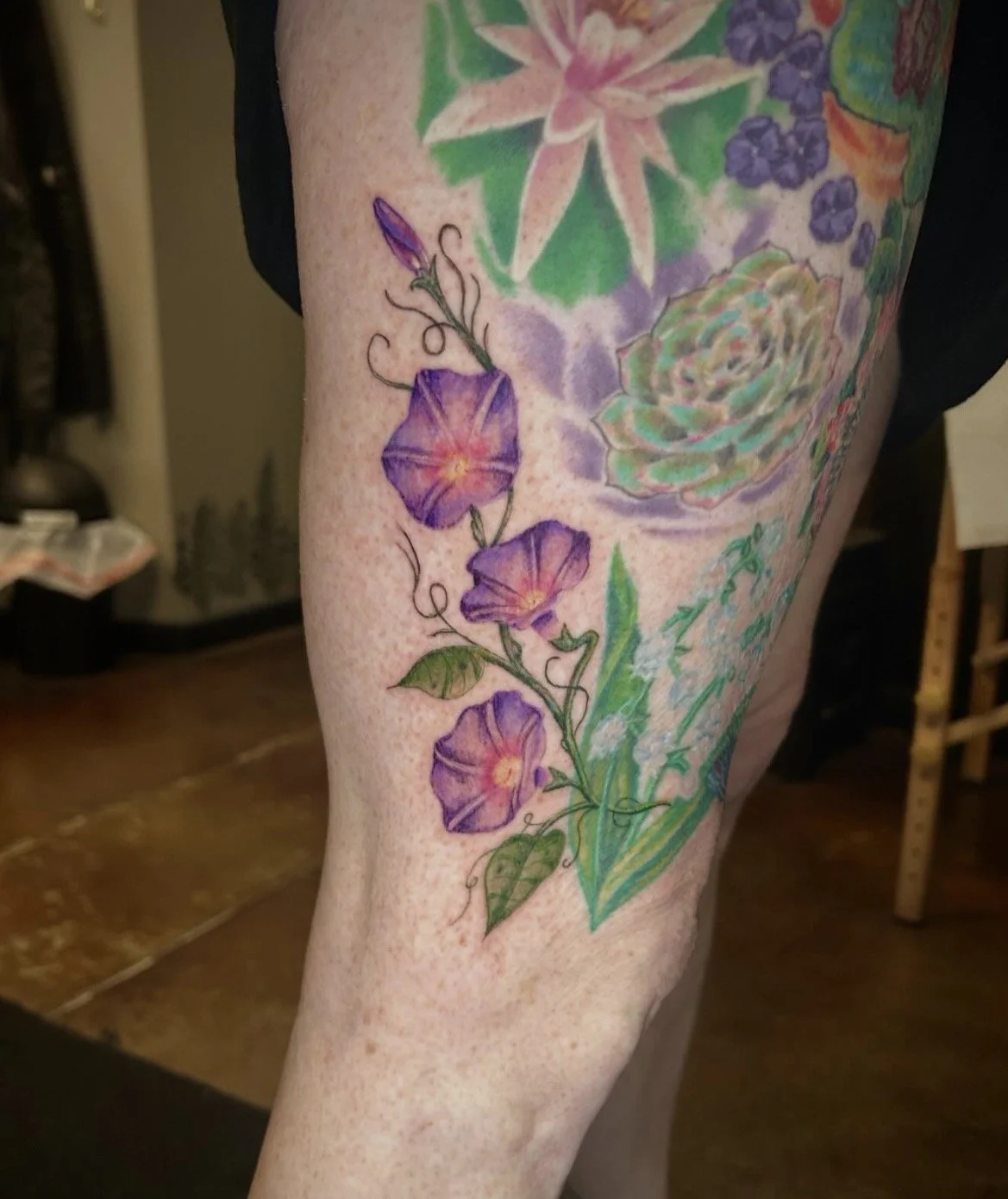 A colorful tattoo on a person's thigh featuring purple morning glories, a rose, and various green leaves with watercolor-style background effects.