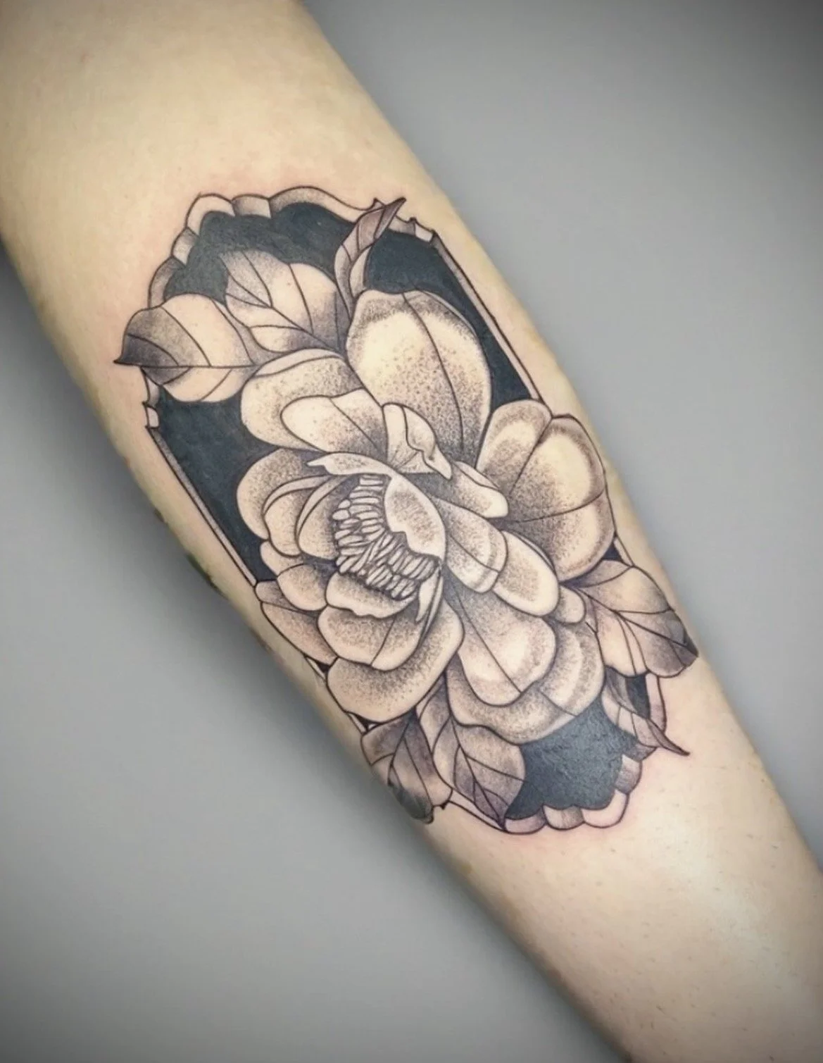 Black and gray floral tattoo of a peony flower and leaves on a person's arm, with a decorative frame.