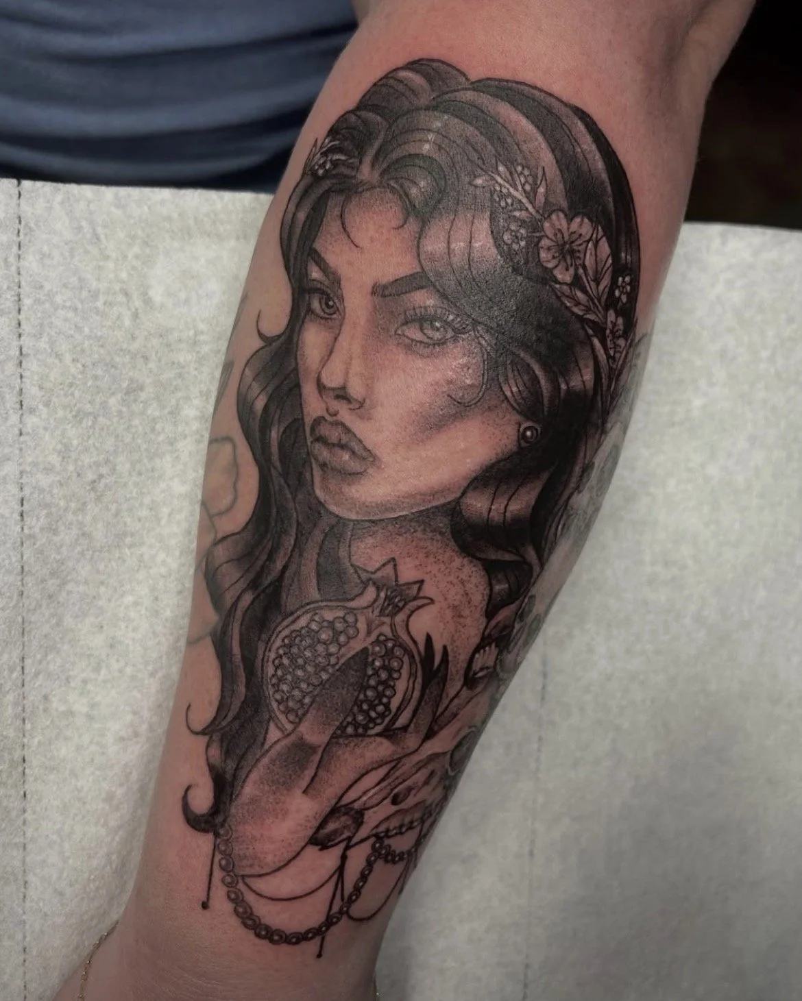 Black and gray tattoo of a woman's face with long wavy hair, flowers, and jewelry on an arm.
