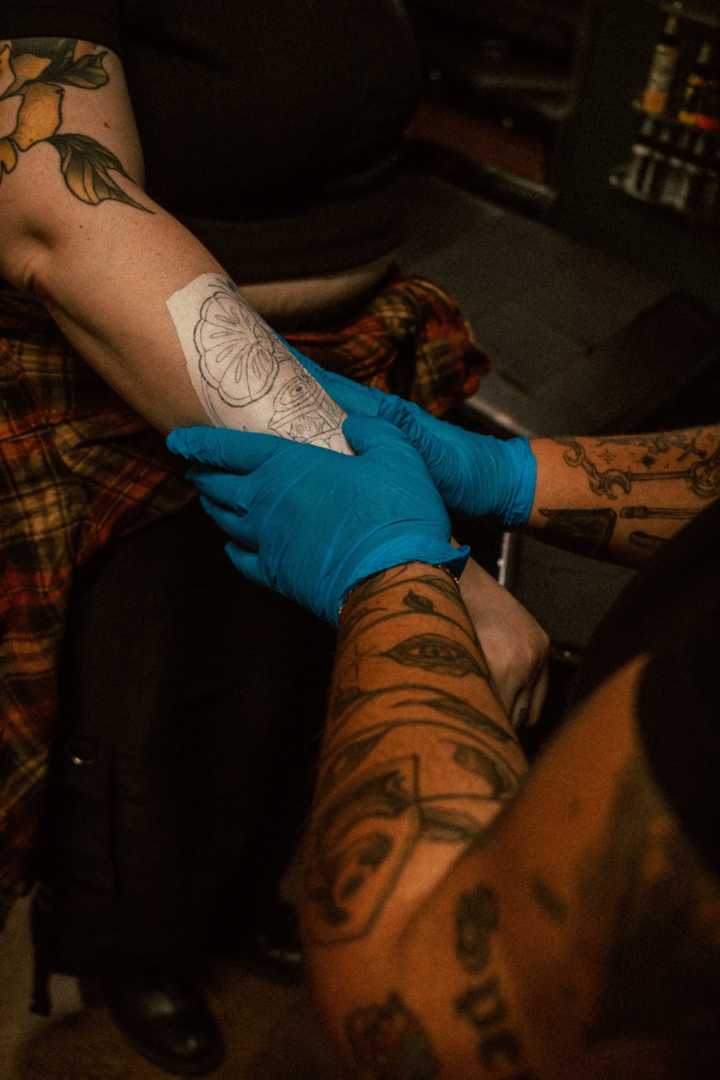 A person with multiple tattoos on their arm and leg is receiving a tattoo, using a tattoo machine, while wearing blue gloves. The background shows a dark room with a bookshelf.