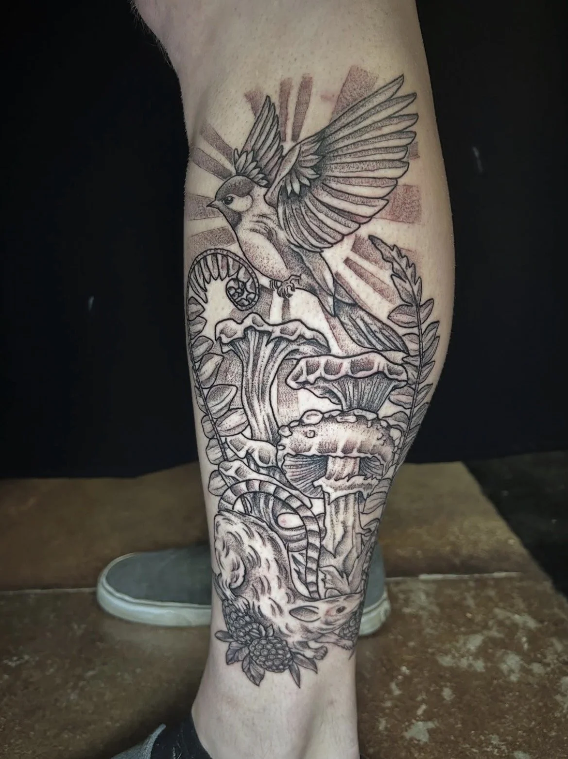 Black and gray tattoo of a bird flying above mushrooms, fern leaves, and a snake wrapped around grapes on a person's leg.