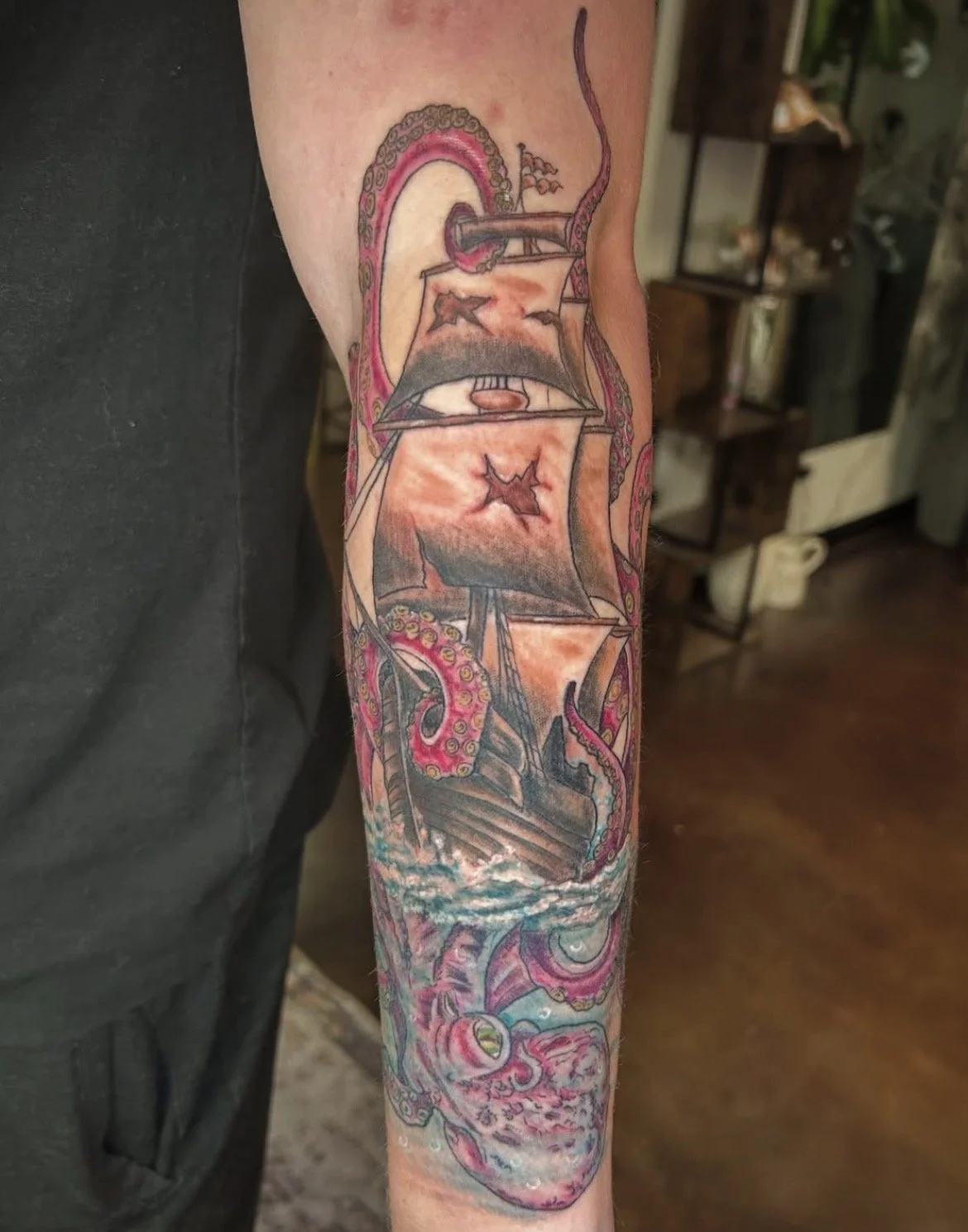 A colorful tattoo on a person's arm depicting a pirate ship engaged in a battle with a tentacled sea creature underground, with the ship firing cannons and the tentacle wrapping around the ship.