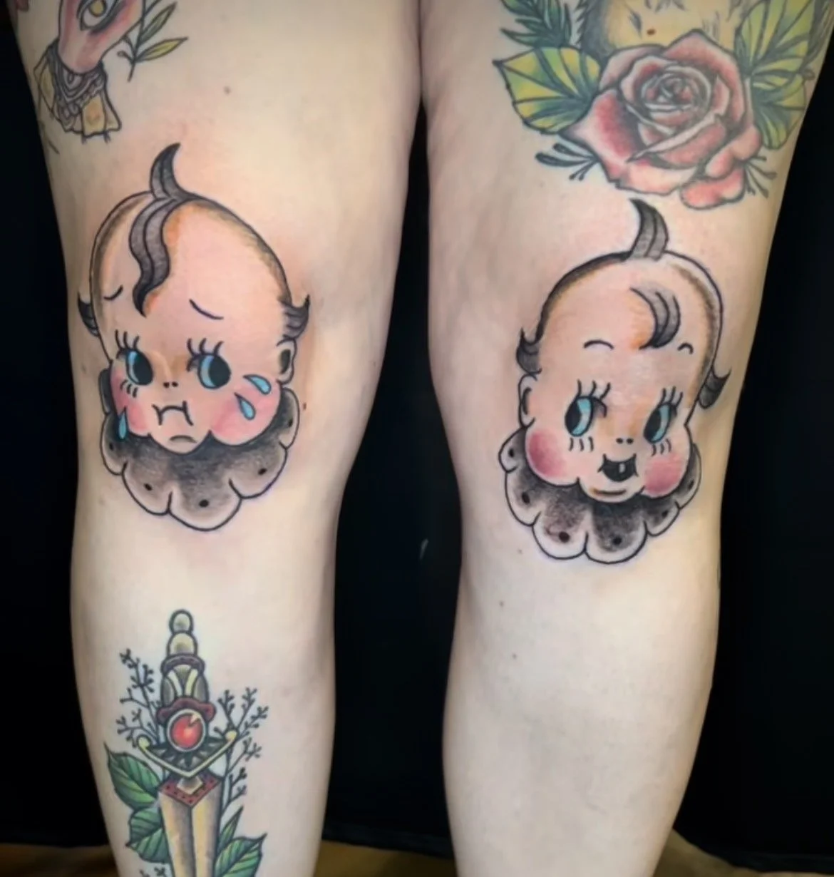 Tattoo of two vintage baby doll faces with rosy cheeks and big eyes covering the front of the thighs, with a rose flower and a decorative dagger design on the lower leg.