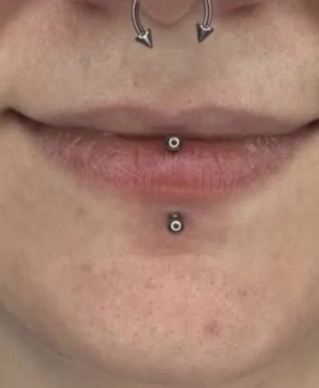 Close-up of a person's lips and chin with two lip piercings and a nose ring visible.