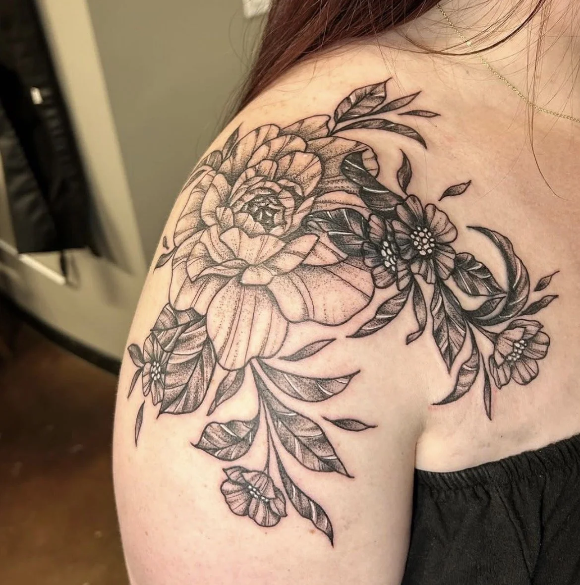 Black and gray floral tattoo featuring large layered peony and smaller flowers with leaves on a person's shoulder.
