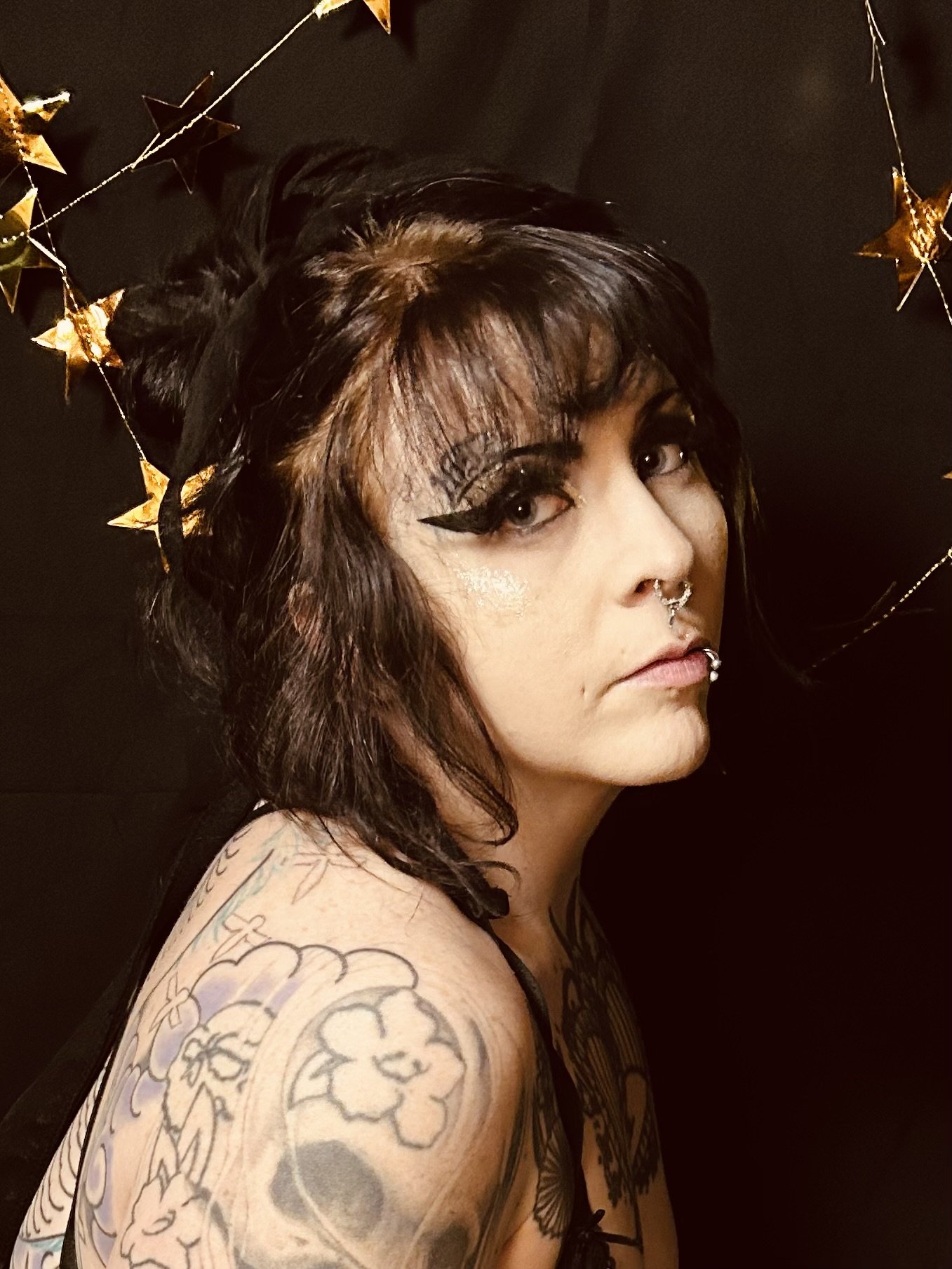 A woman with dark, wavy hair and multiple tattoos on her shoulder and arm, wearing dramatic eye makeup, piercings on her nose and lips, and a black tank top. She is standing against a dark background with a gold star garland.