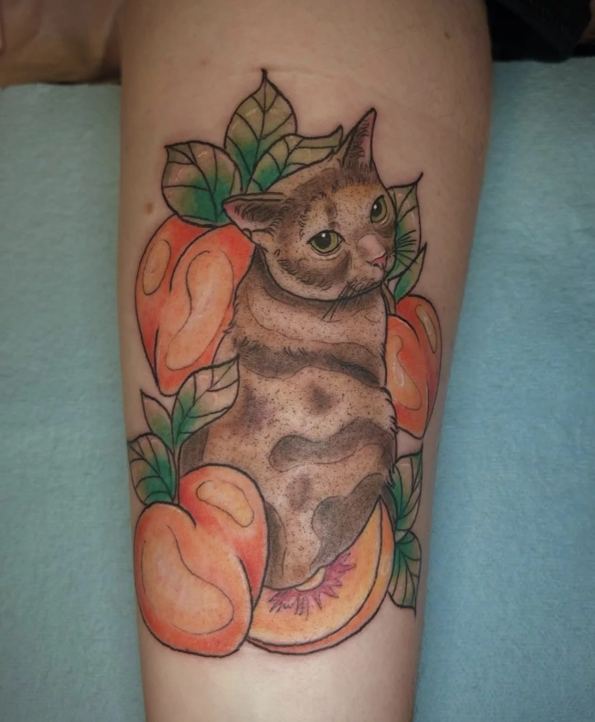 Tattoo of a cat's face surrounded by a peach and green leaves, with some peaches around it.