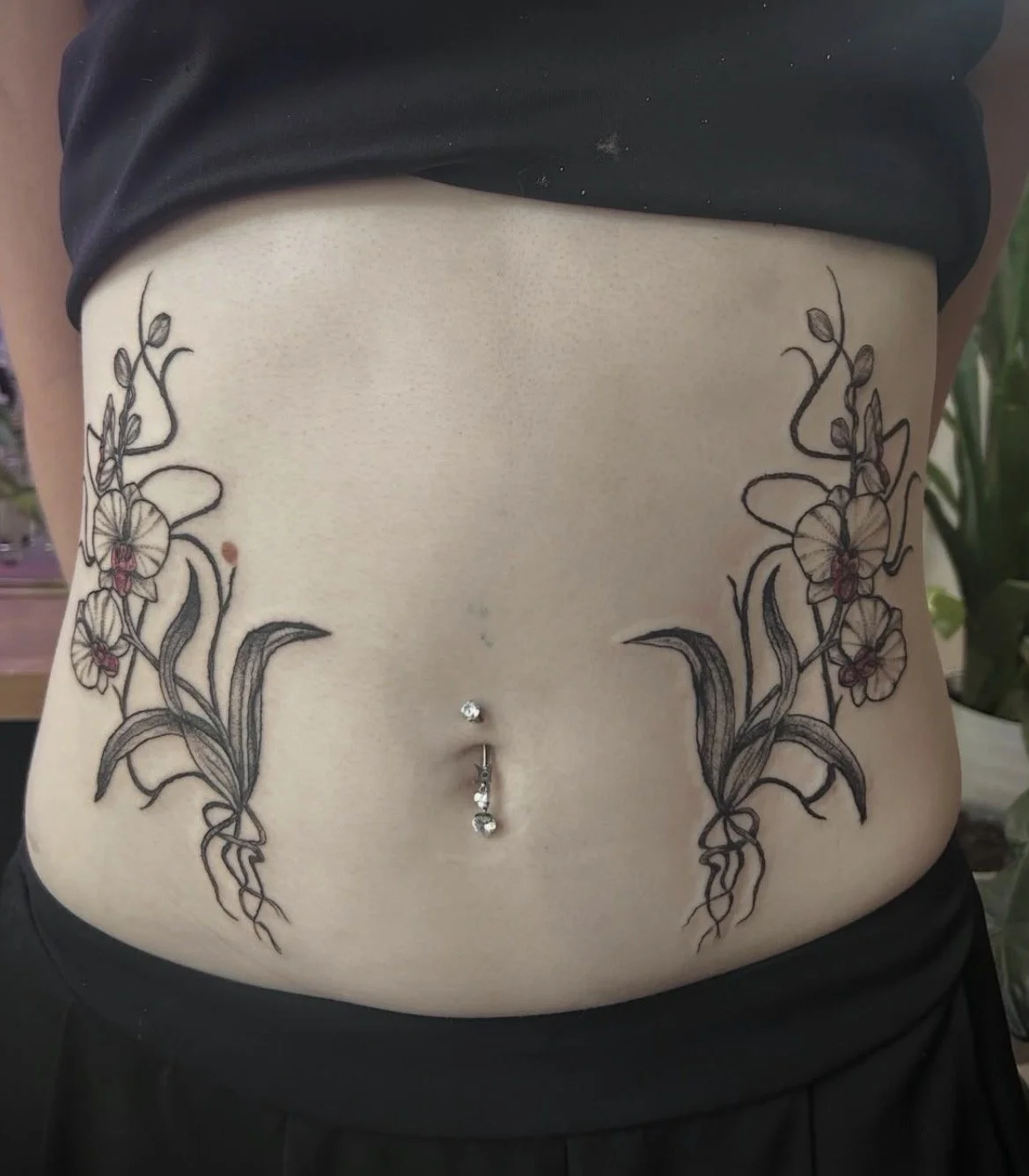 Tattoo of floral vines with orchids on each side of a person's lower abdomen, with a belly button piercing and navel jewelry.
