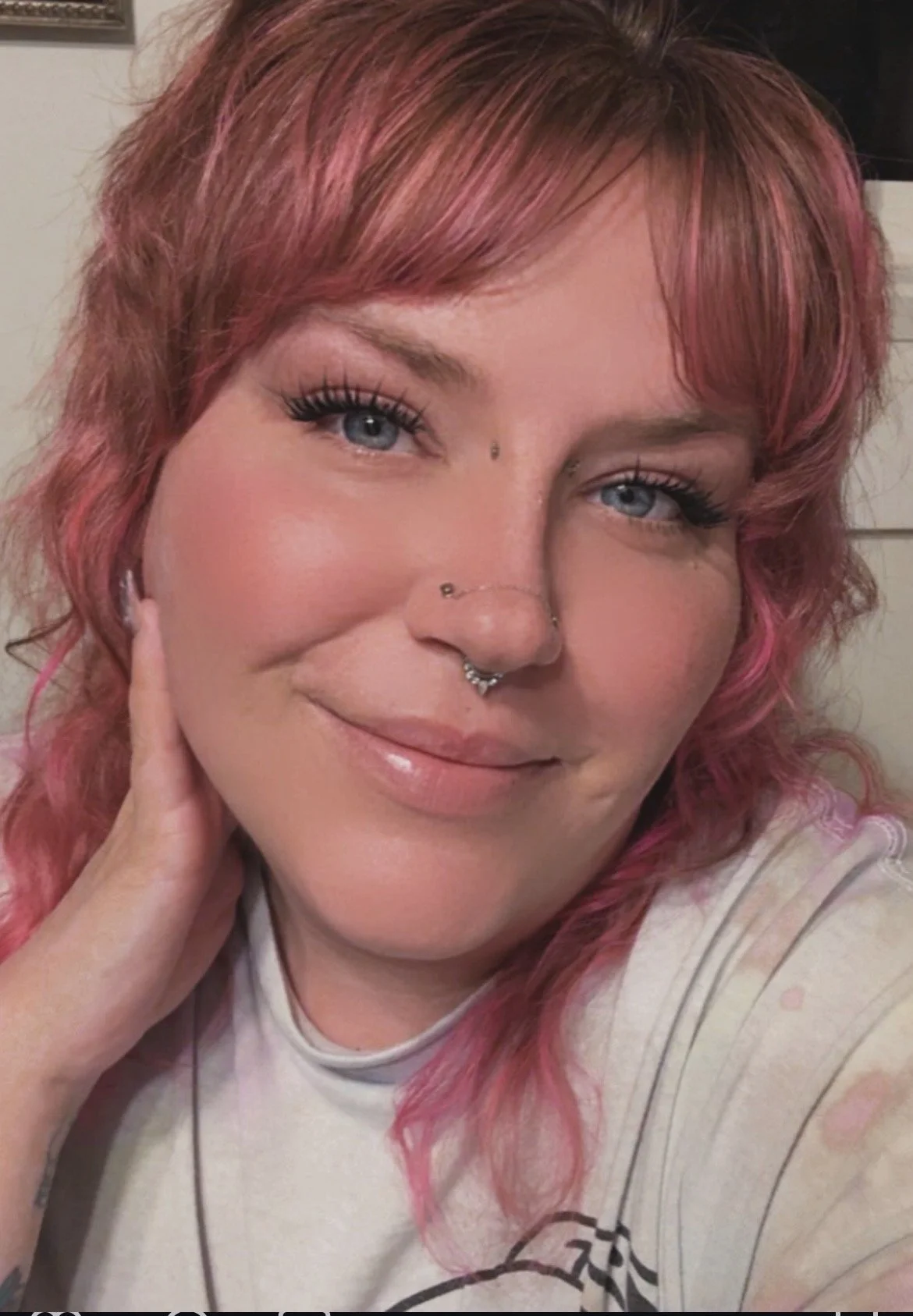 A woman with pink hair, blue eyes, and multiple facial piercings wearing makeup and a white shirt, smiling at the camera, resting her hand on her face.