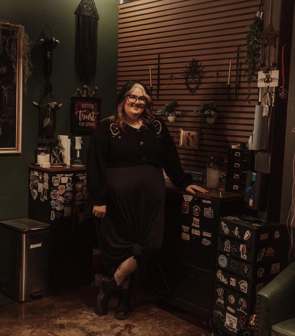 A woman with glasses and a smile, dressed in a black outfit, stands in a dimly lit room decorated with wall art, plants, and stickers on furniture, next to a small cabinet and a desk.