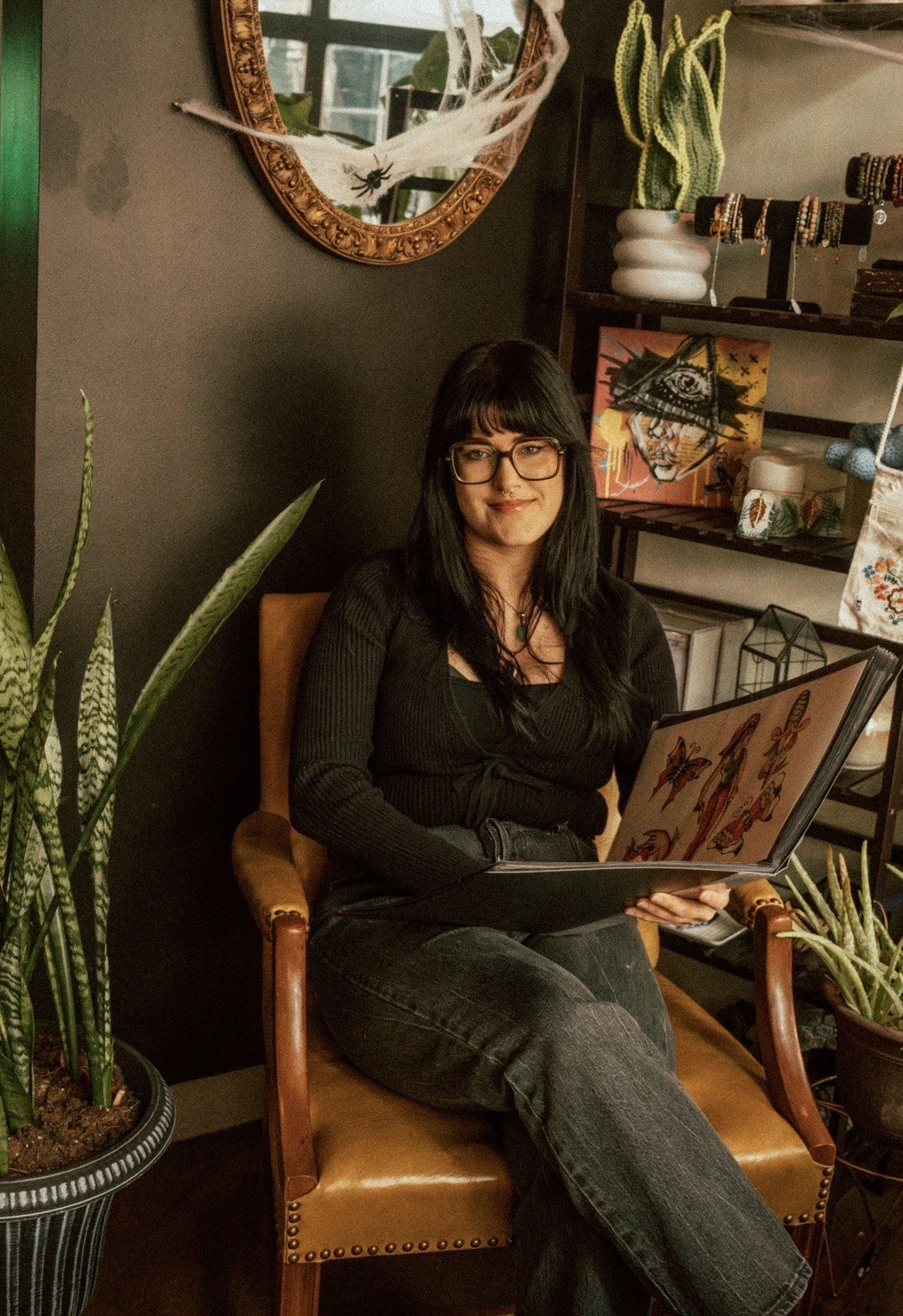 A woman with long dark hair, glasses, and a black shirt sits in a wooden chair holding a menu with butterfly illustrations. She is in a cozy room with plants, artwork, and shelves decorated with jewelry and art supplies.