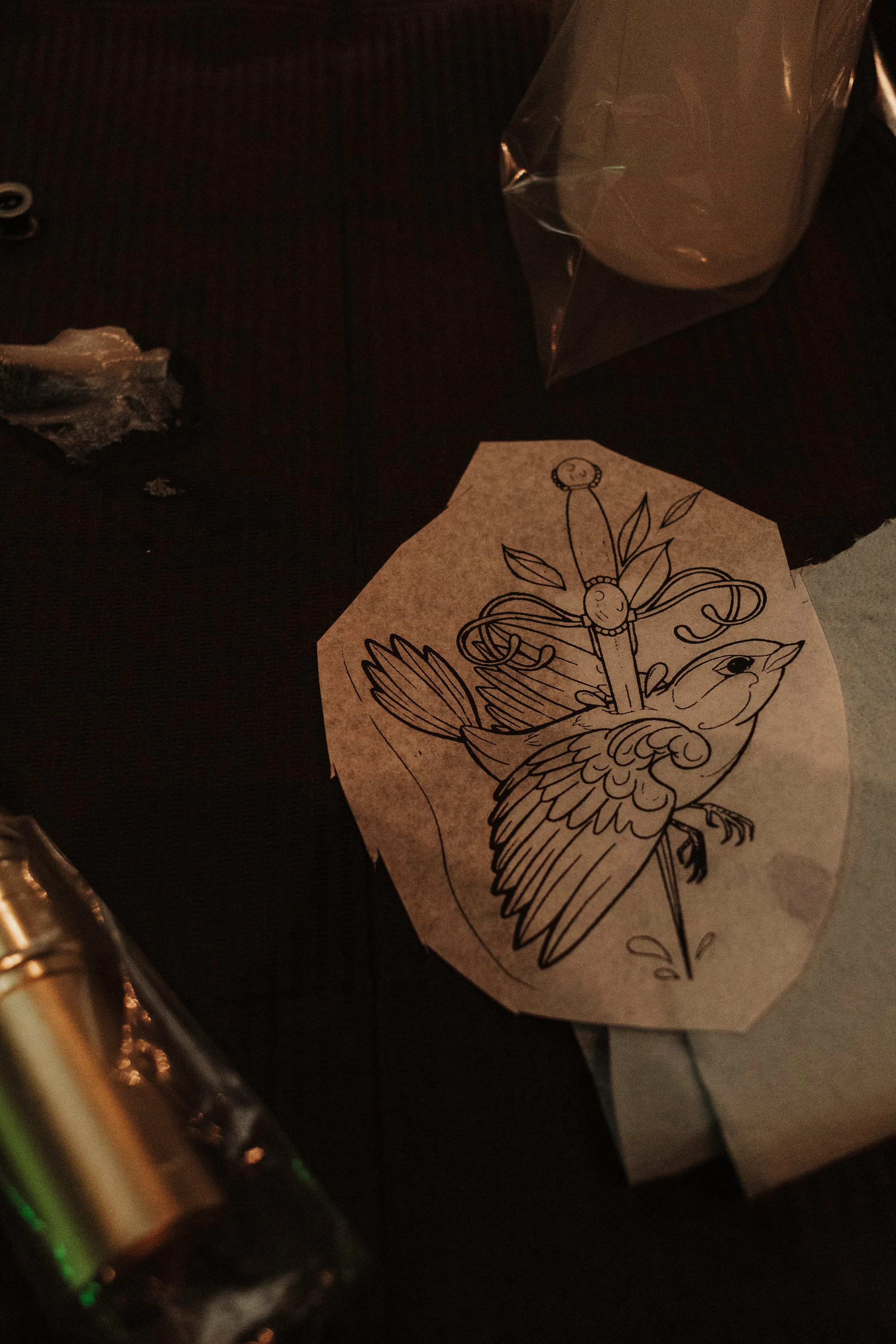 A hand-drawn coloring page featuring a bird with a sword surrounded by decorative leaves and scrolls, placed on a dark surface with a plastic-wrapped package and a piece of meat nearby.