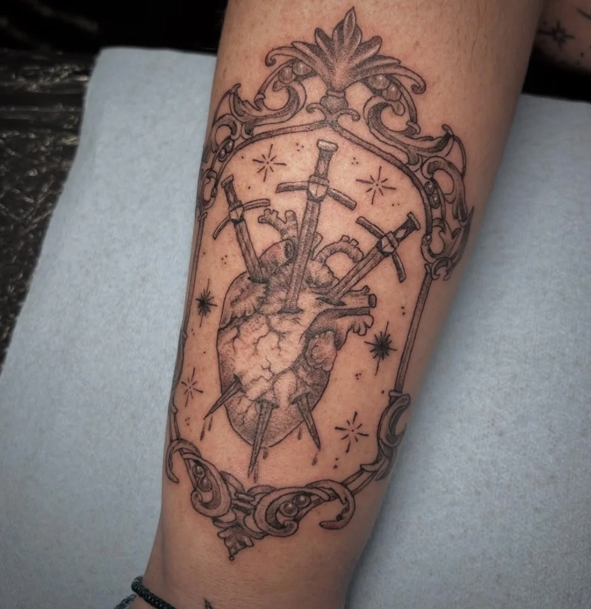 Tattoo of an anatomical heart pierced by multiple swords, framed with ornate decorative elements and surrounded by small star-like symbols.
