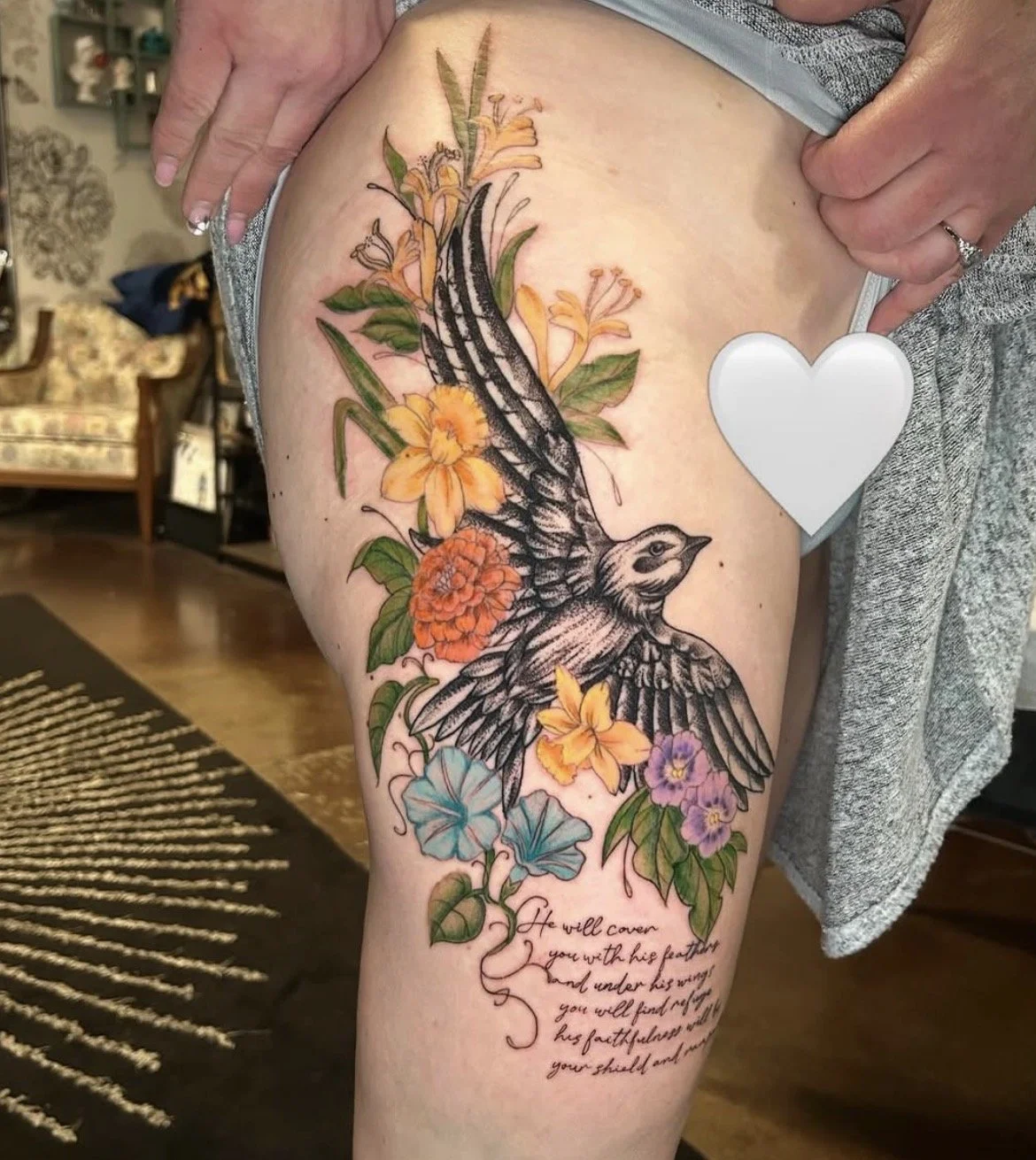 A colorful tattoo of a bird with outstretched wings surrounded by orange, yellow, purple, and blue flowers and green leaves on a person's thigh. There is a handwritten quote underneath the tattoo.