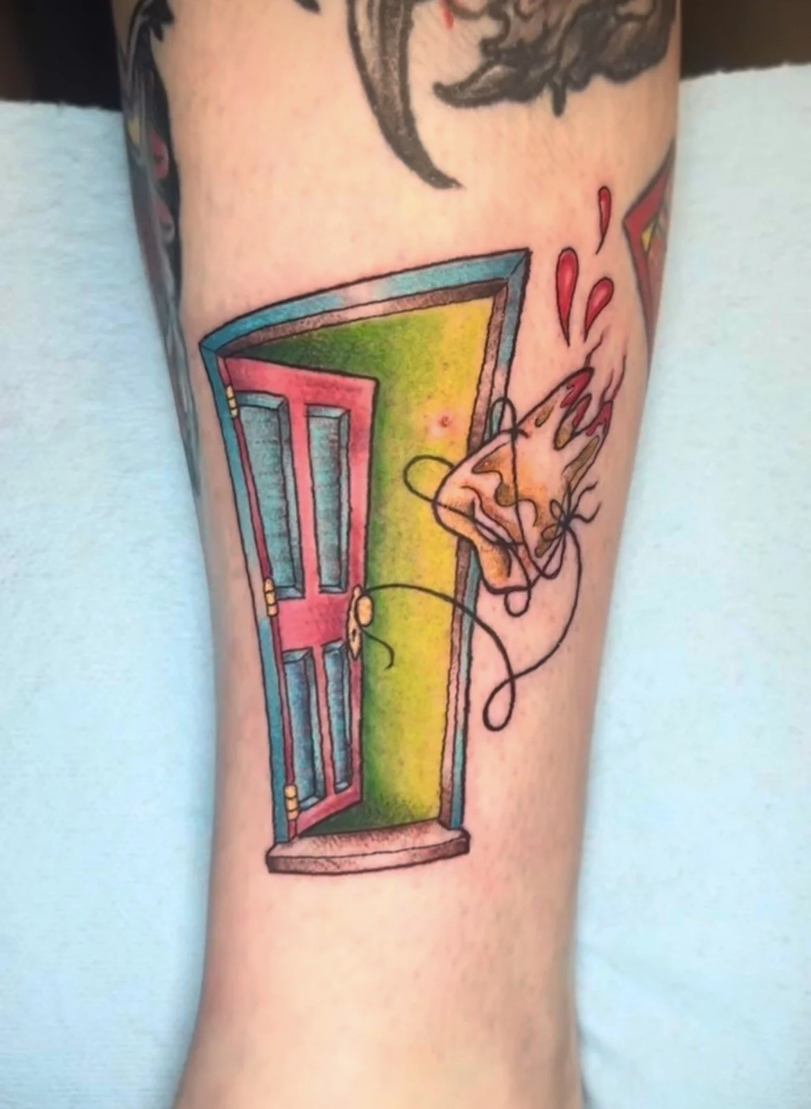 Tattoo of a colorful door (pink, blue, green) with a black outline, slightly ajar, on a person's arm. The door appears to be opening to a rainbow-colored background. The tattoo includes a line drawing of a hand holding a string attached to a ball, wi