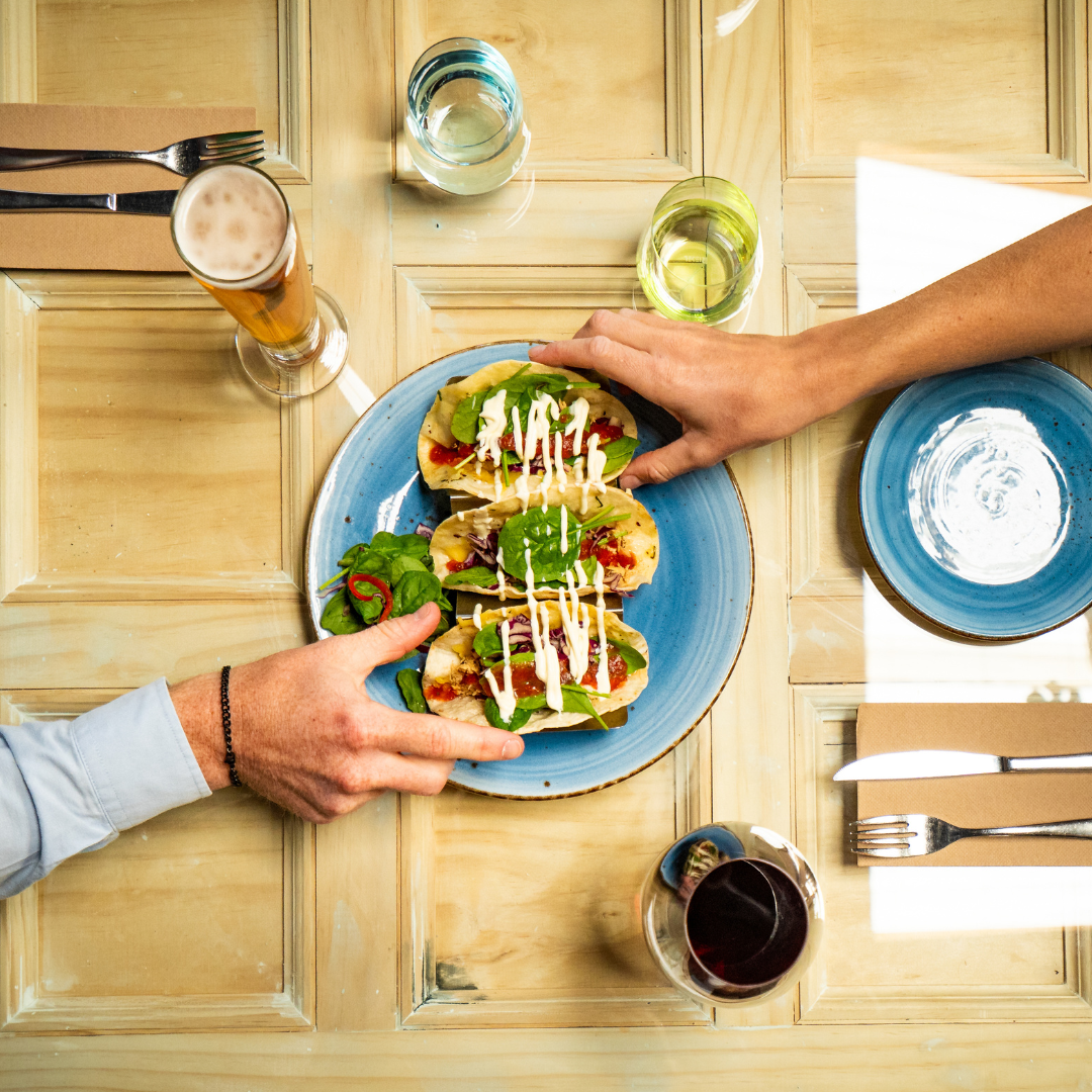 Two hands reaching for tacos on a blue plate at a wooden table with drinks and utensils.