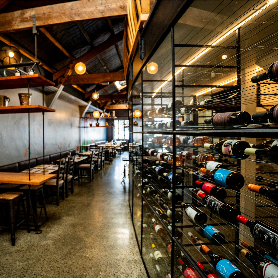 Interior of a wine restaurant with tables and chairs on the left and a glass wine rack filled with wine bottles on the right, wooden ceiling, warm lighting, and a rustic ambiance.