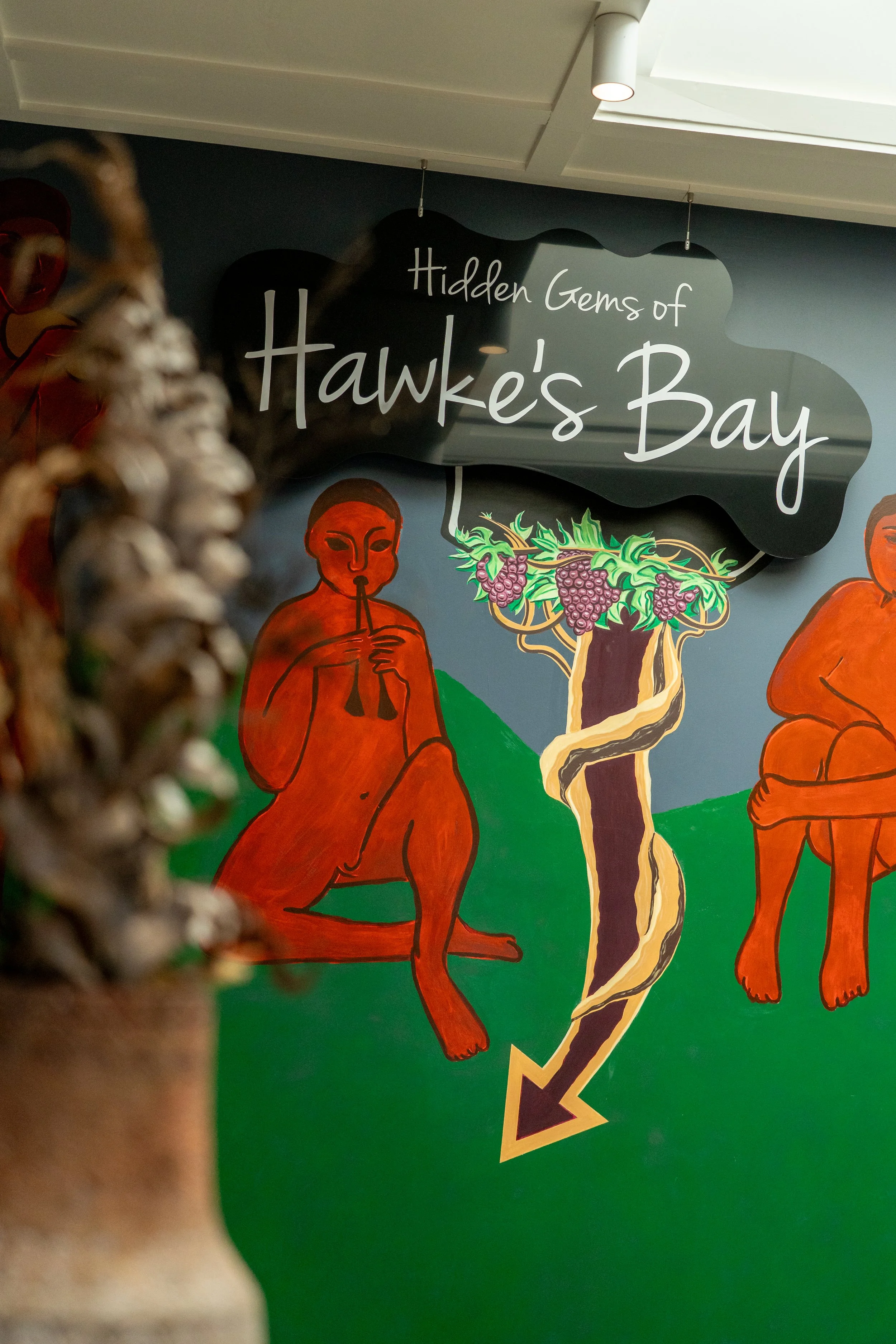 Wall sign reading 'Hidden Gems of Hawke's Bay' with colorful illustrated figures and a grapevine, featuring a person playing a flute.